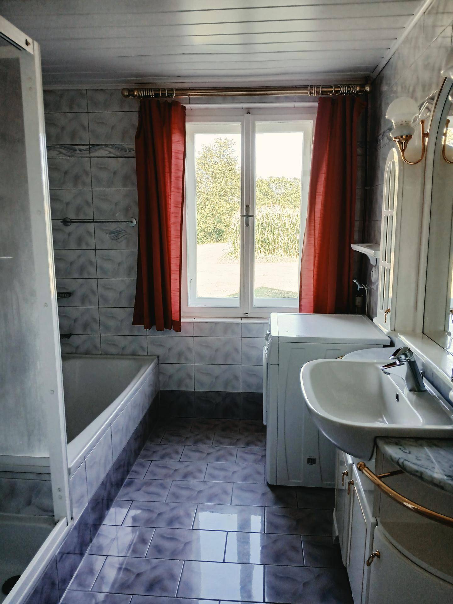 Gefliestes Badezimmer mit Badewanne, Dusche, Waschbecken und Fenster mit Blick ins Grüne.