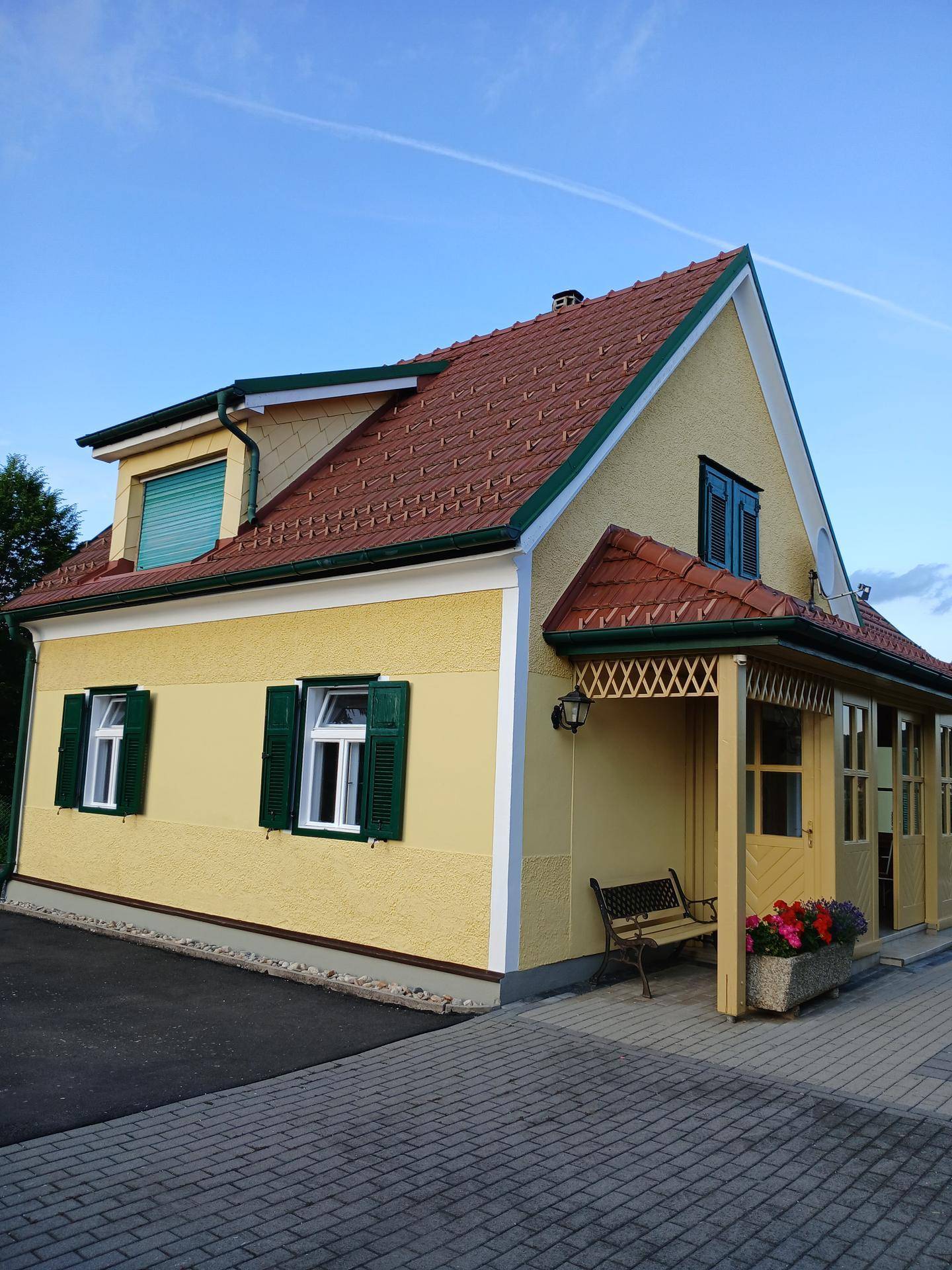 Seitenansicht des gelben Hauses mit roten Dachziegeln, grünen Fensterläden und überdachtem Eingang.