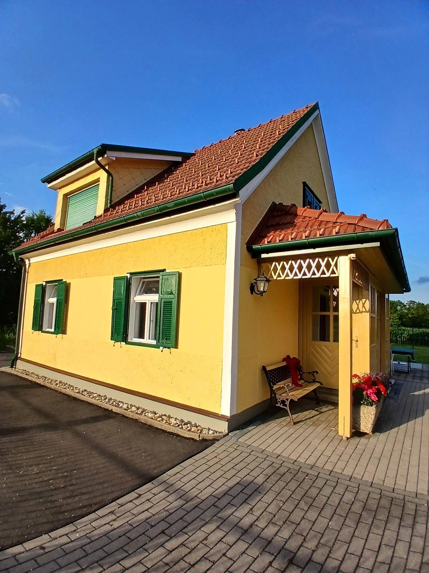 Gelbes Haus mit rotem Ziegeldach, grünen Fensterläden und einem einladenden überdachten Eingangsbereich.