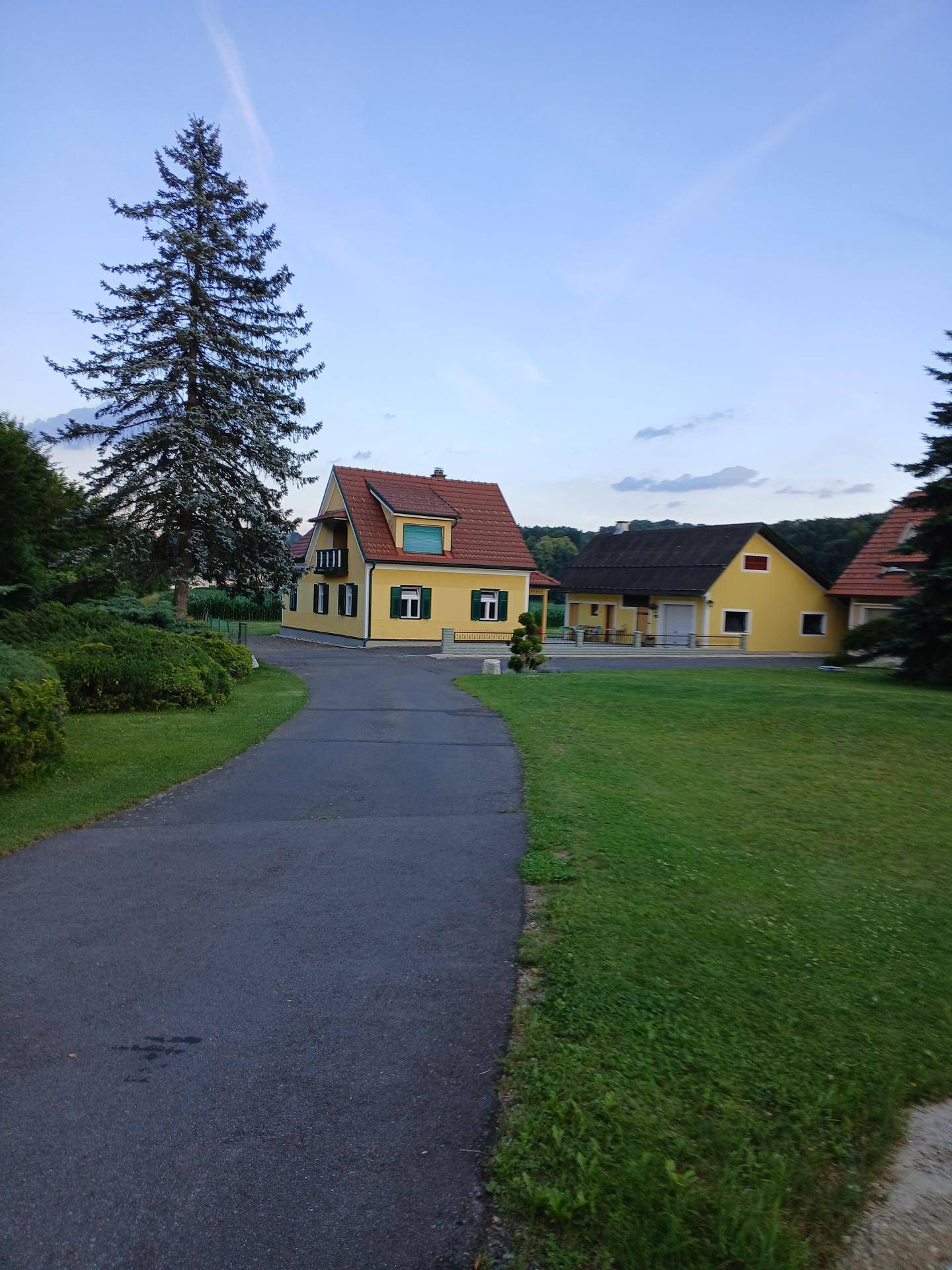 Lange Auffahrt führt zu einem gelben Haus mit rotem Dach und hohen Bäumen in ländlicher Umgebung.
