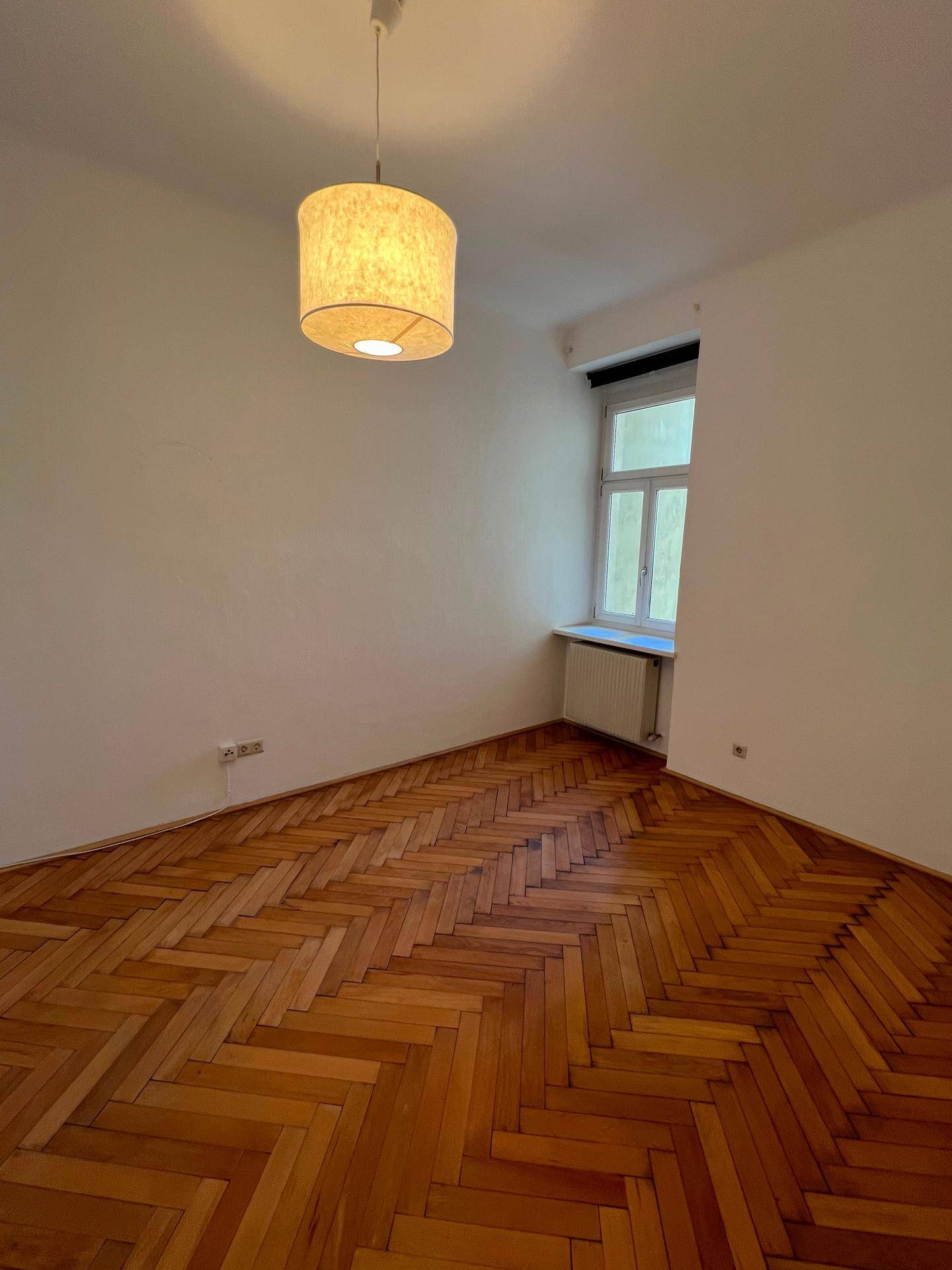 Helles Zimmer mit Fischgrätparkett und einem Fenster, das Tageslicht spendet.