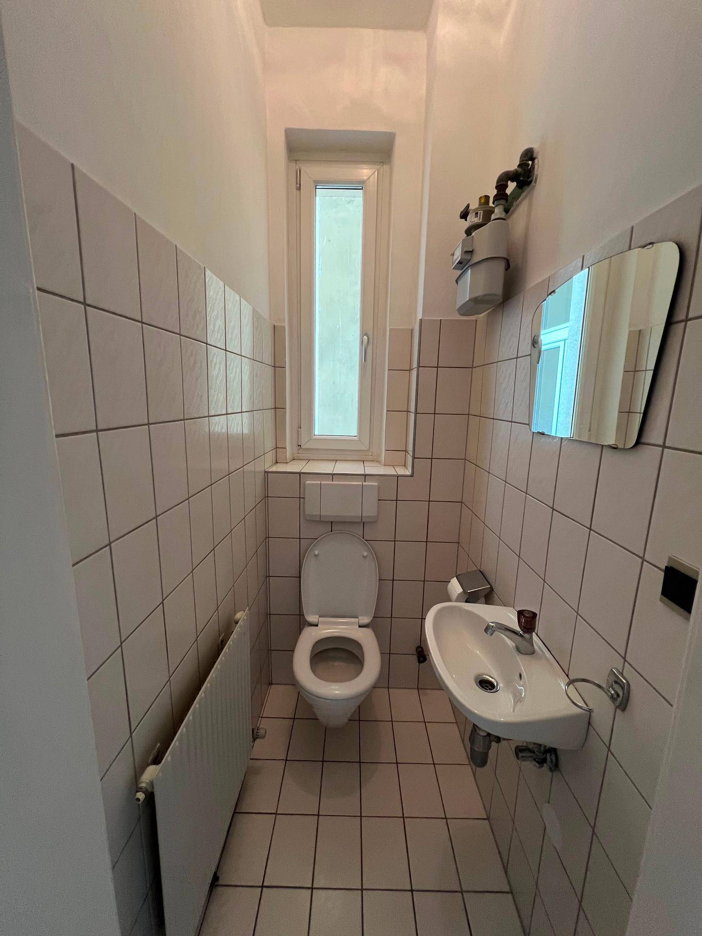 Separates WC mit Fenster, Toilette und kleinem Waschbecken, gefliest in hellem Rosa.