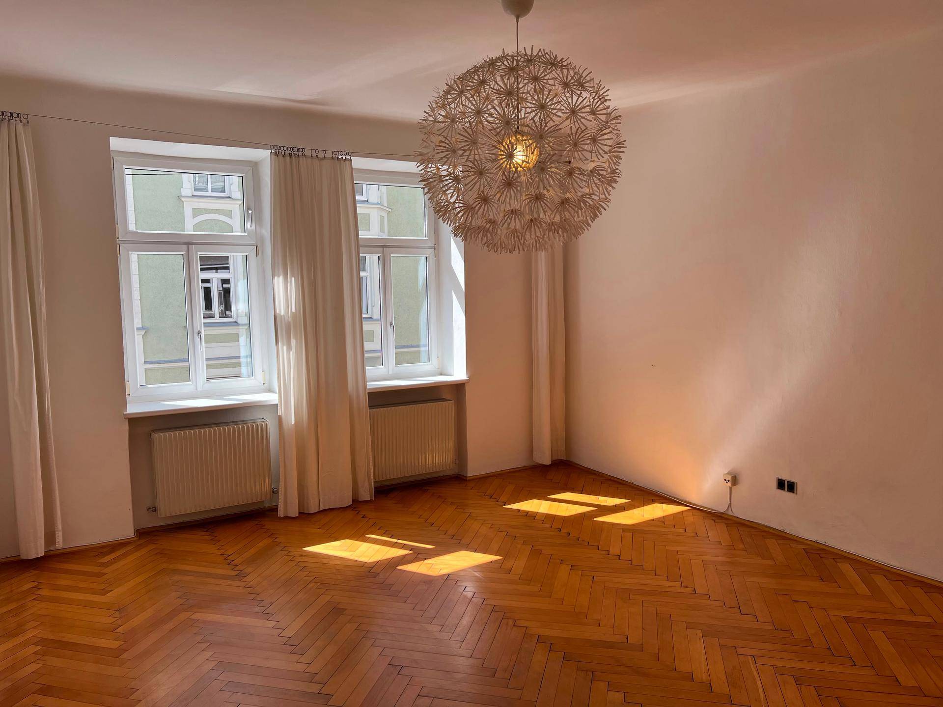 Geräumiges Wohnzimmer mit Fischgrätparkett und großen Fenstern, die viel Licht hereinlassen.