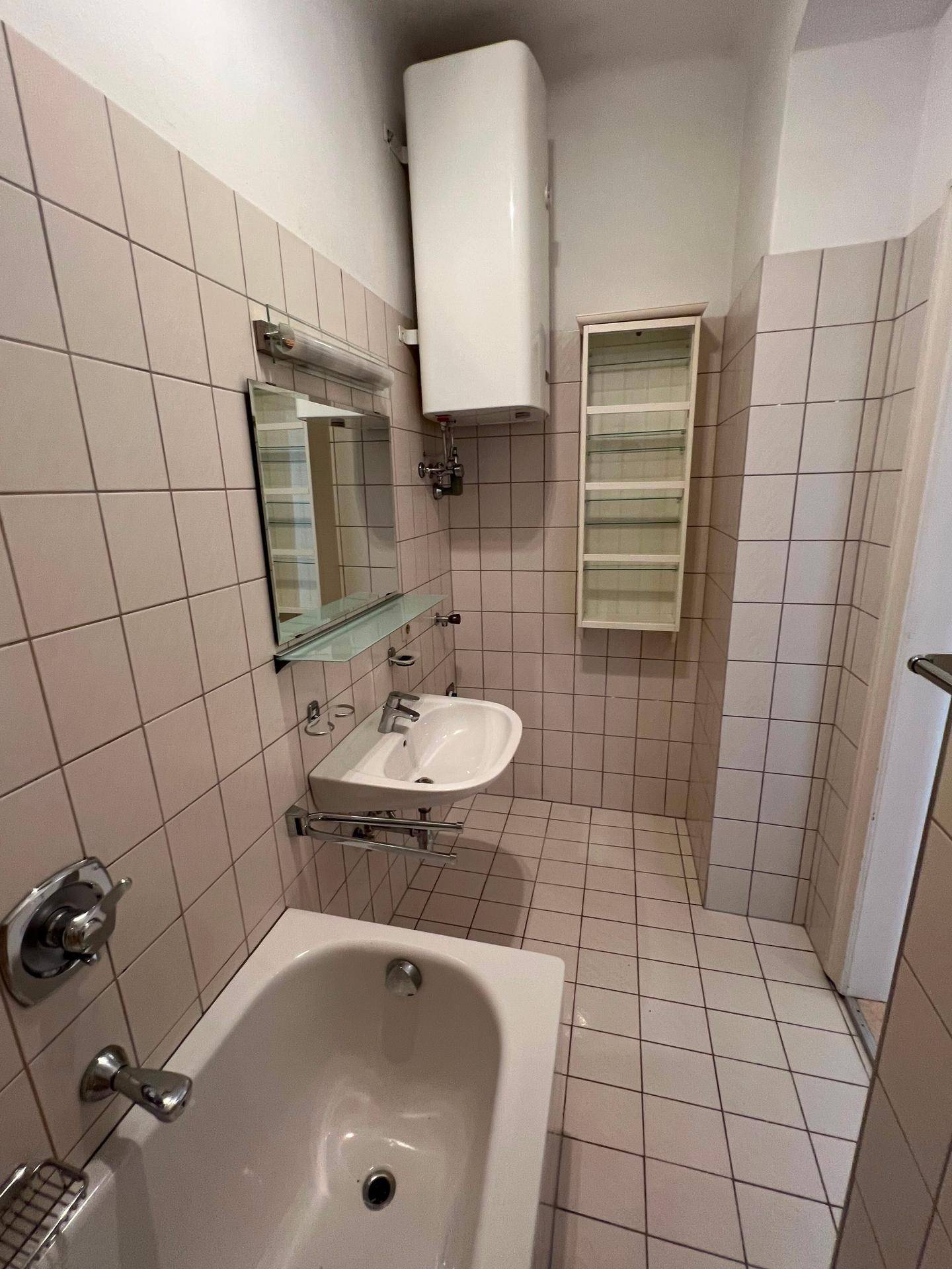 Badezimmer mit Badewanne, Waschbecken und hellrosa Fliesen, ausgestattet mit einem Boiler.