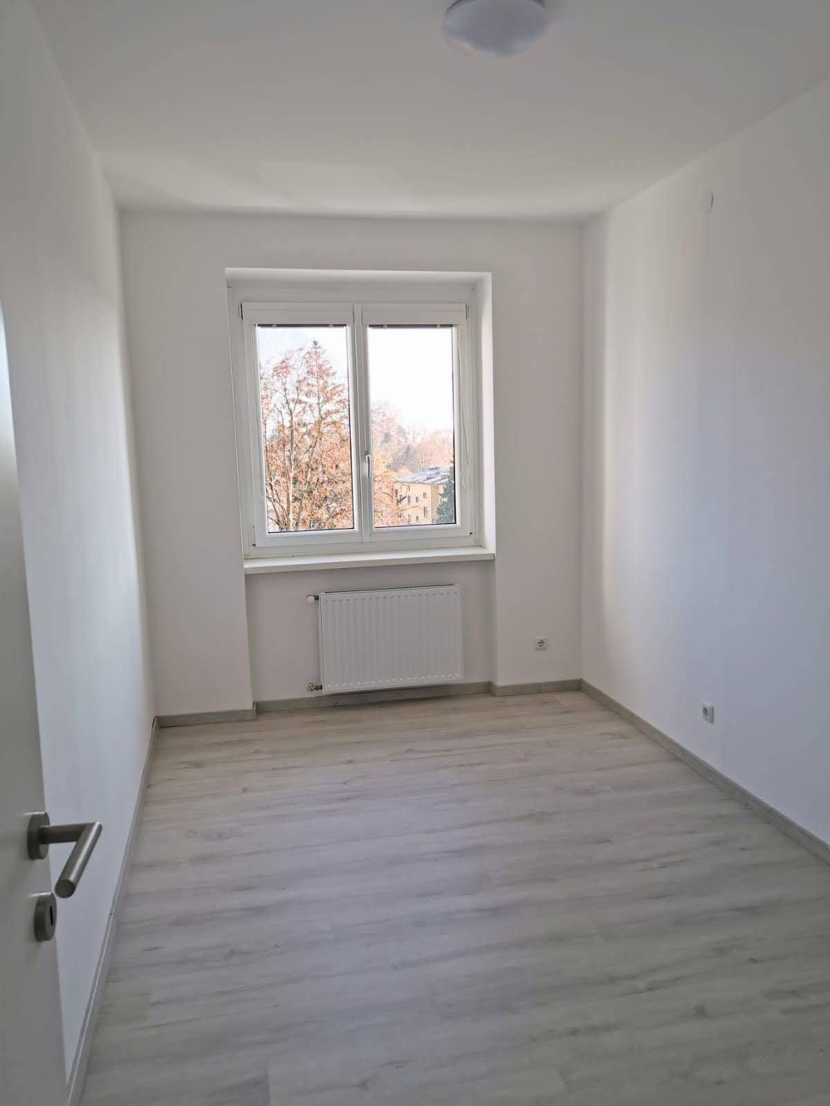 Leeres Schlafzimmer mit hellem Laminatboden und einem Fenster, das viel Tageslicht hereinlässt.