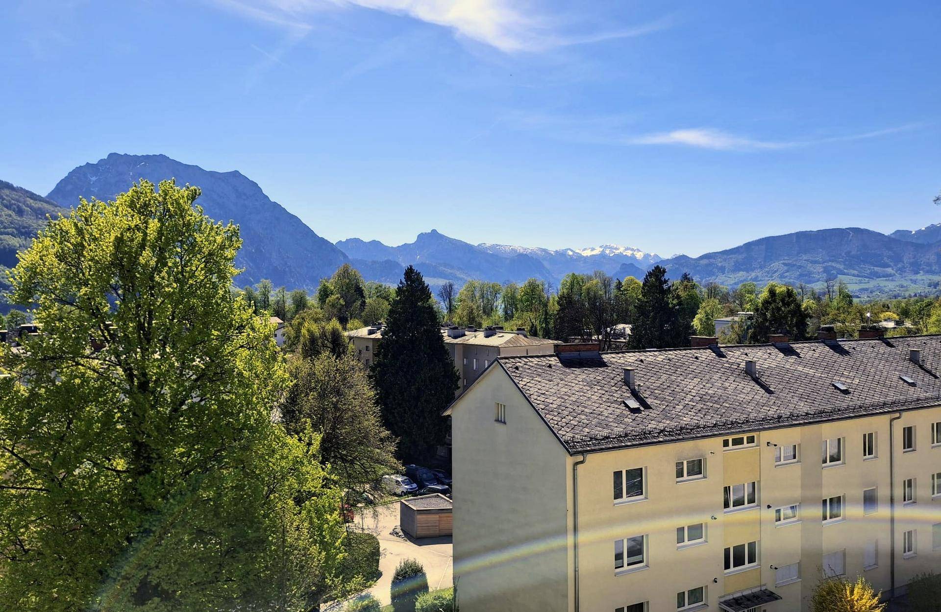 Weitläufiger Ausblick auf schneebedeckte Berge und grüne Hügel mit Wohnbebauung im Vordergrund.