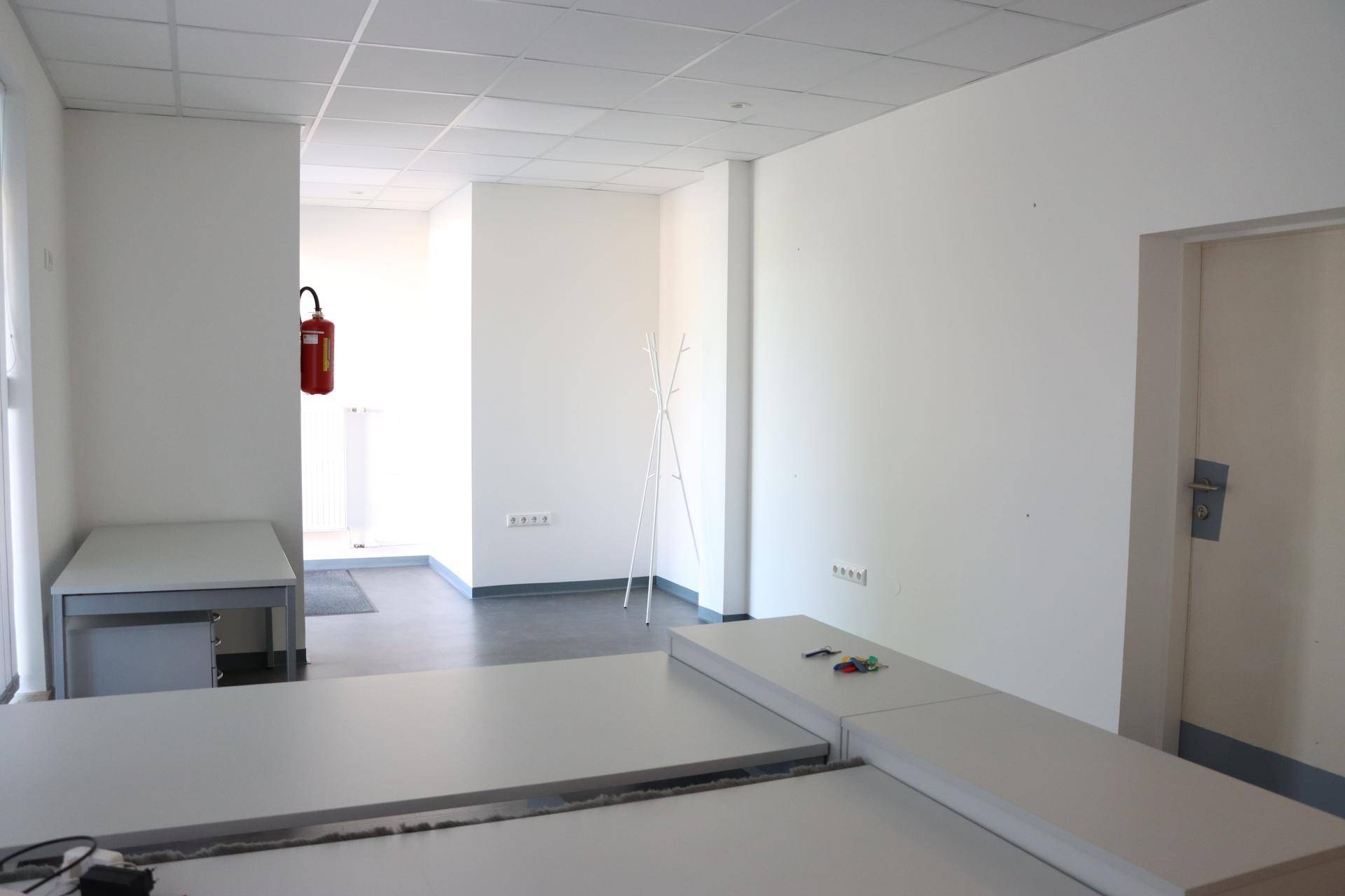 Heller Büroraum mit weißen Wänden, modularen Tischen und Zugang zu einem weiteren Arbeitsbereich.