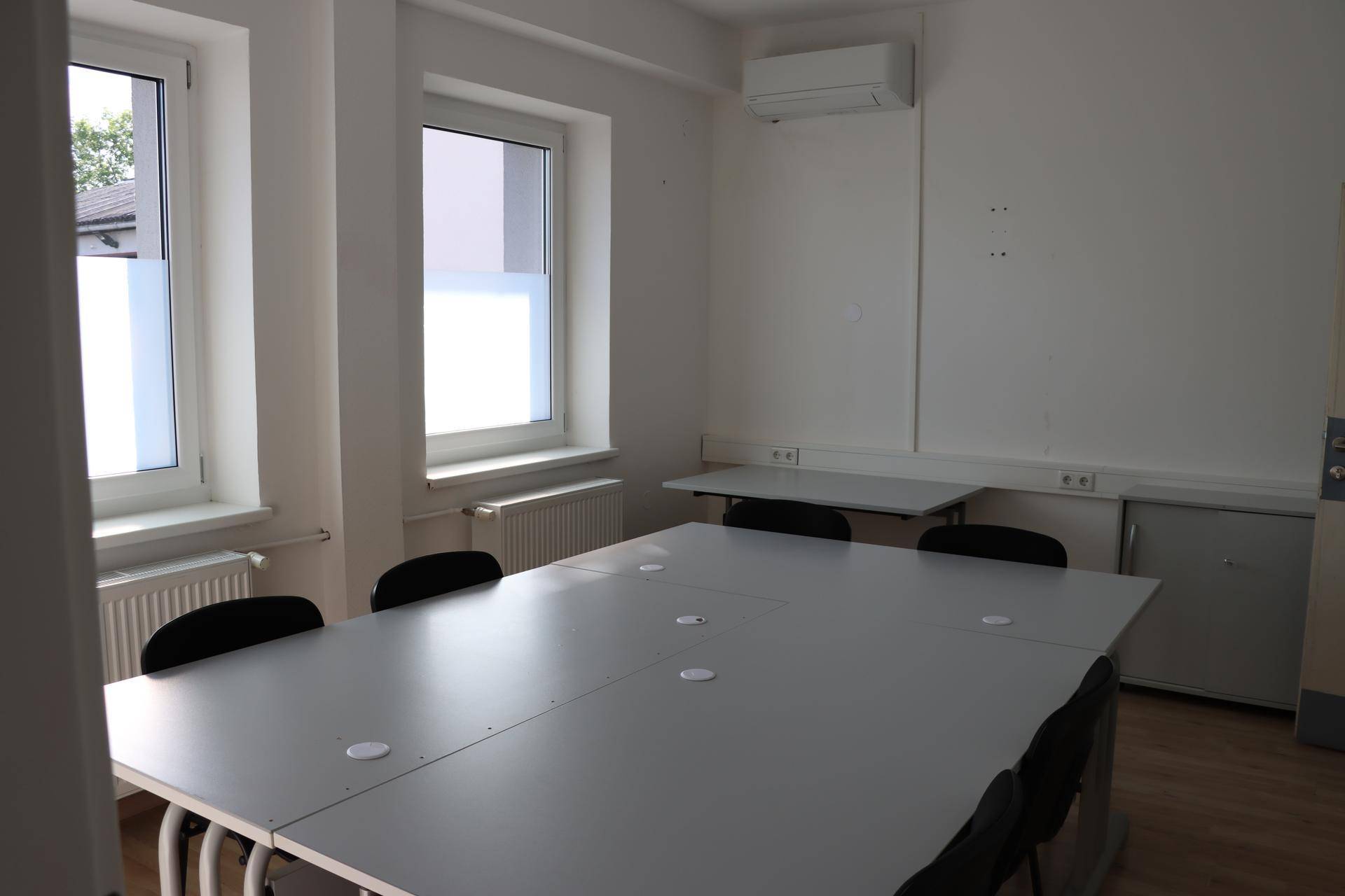 Heller Konferenzraum mit zwei Fenstern, Klimaanlage und einem großen modularen Tisch für Besprechungen.