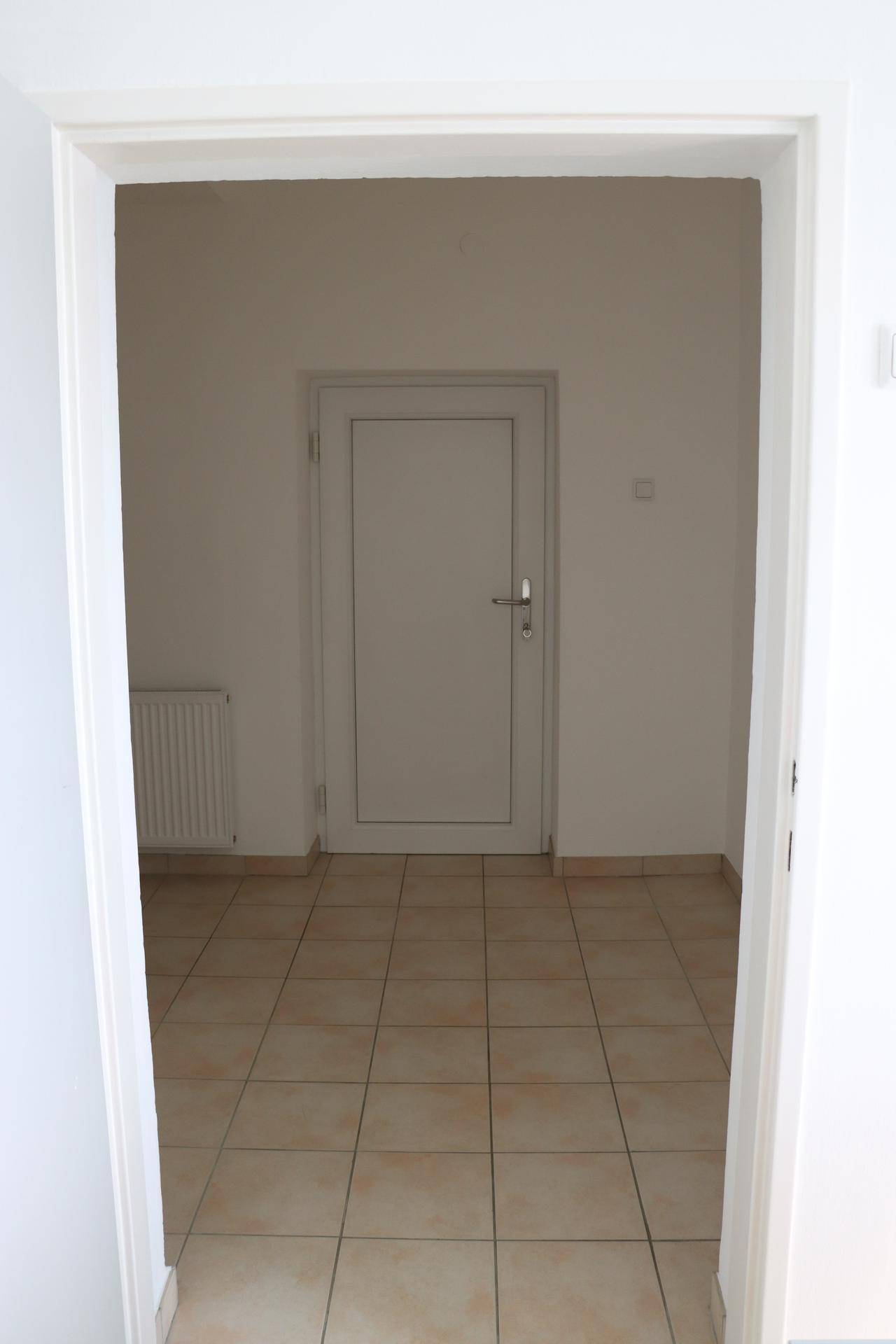 Heller Flur mit beigefarbenen Fliesen, weißer Tür und einem Heizkörper an der Wand.