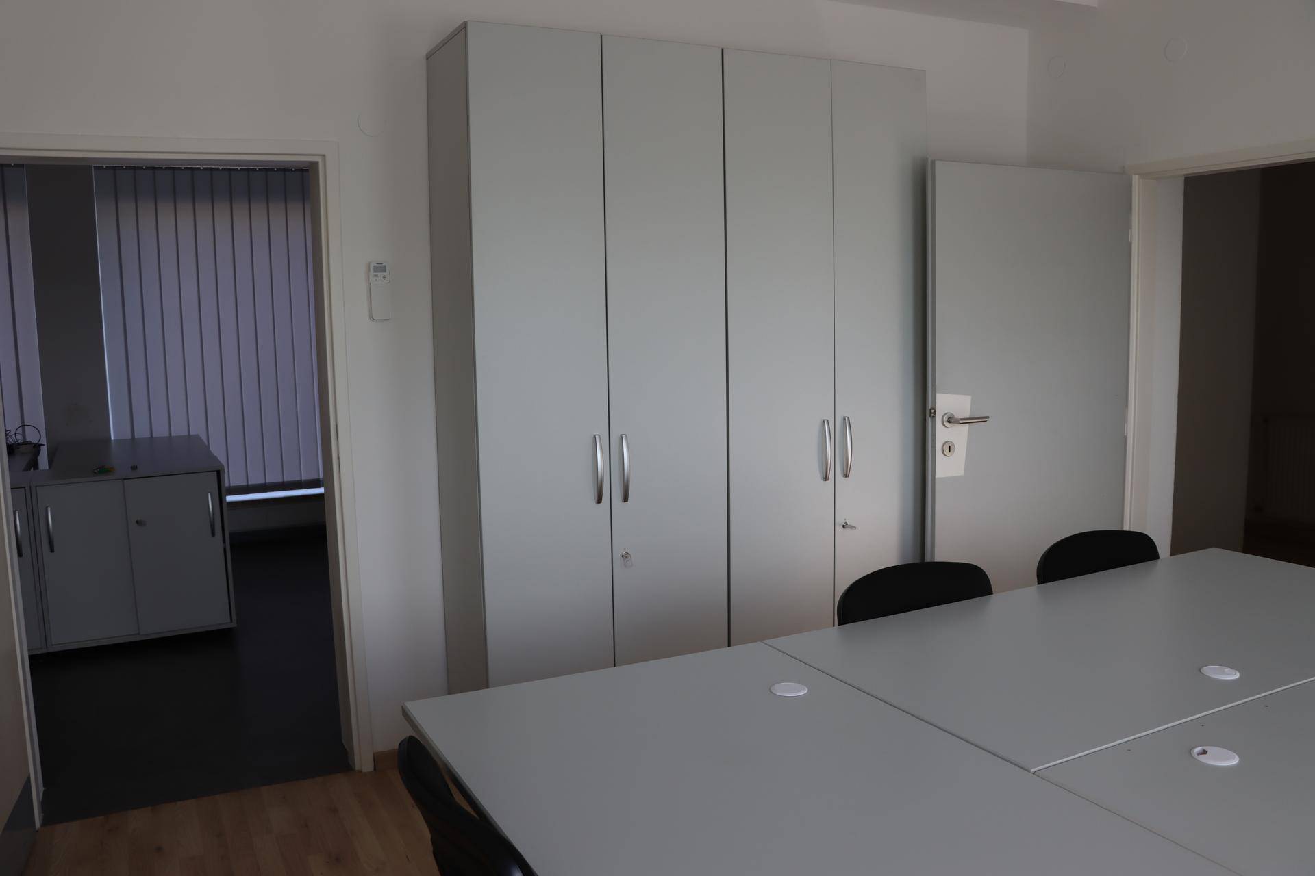 Konferenzraum mit grauen Schränken, großem Tisch und Zugang zu einem weiteren Bürobereich.