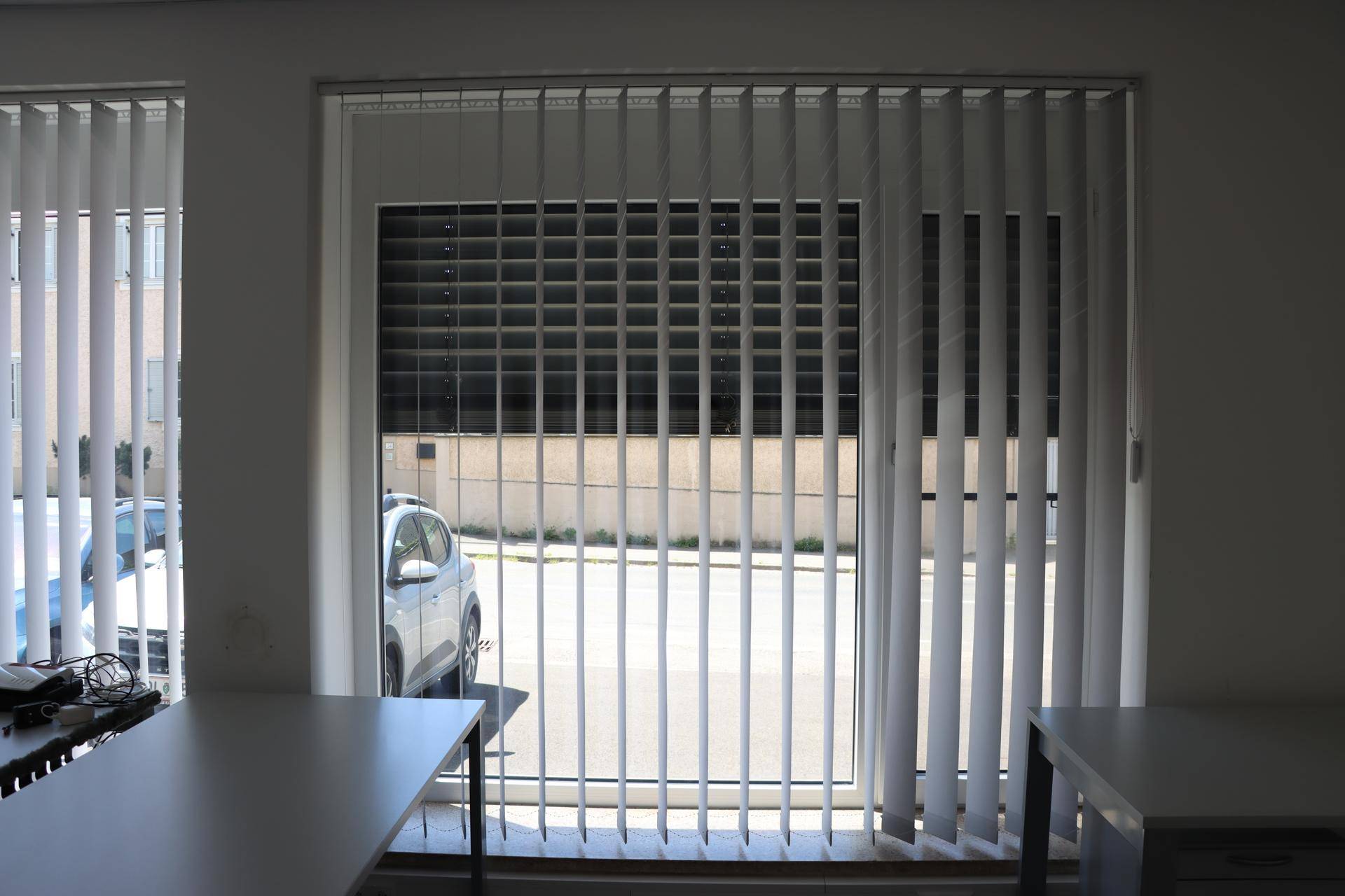 Großes Fenster mit vertikalen Jalousien, das den Blick auf die Straße und ein geparktes Auto freigibt.