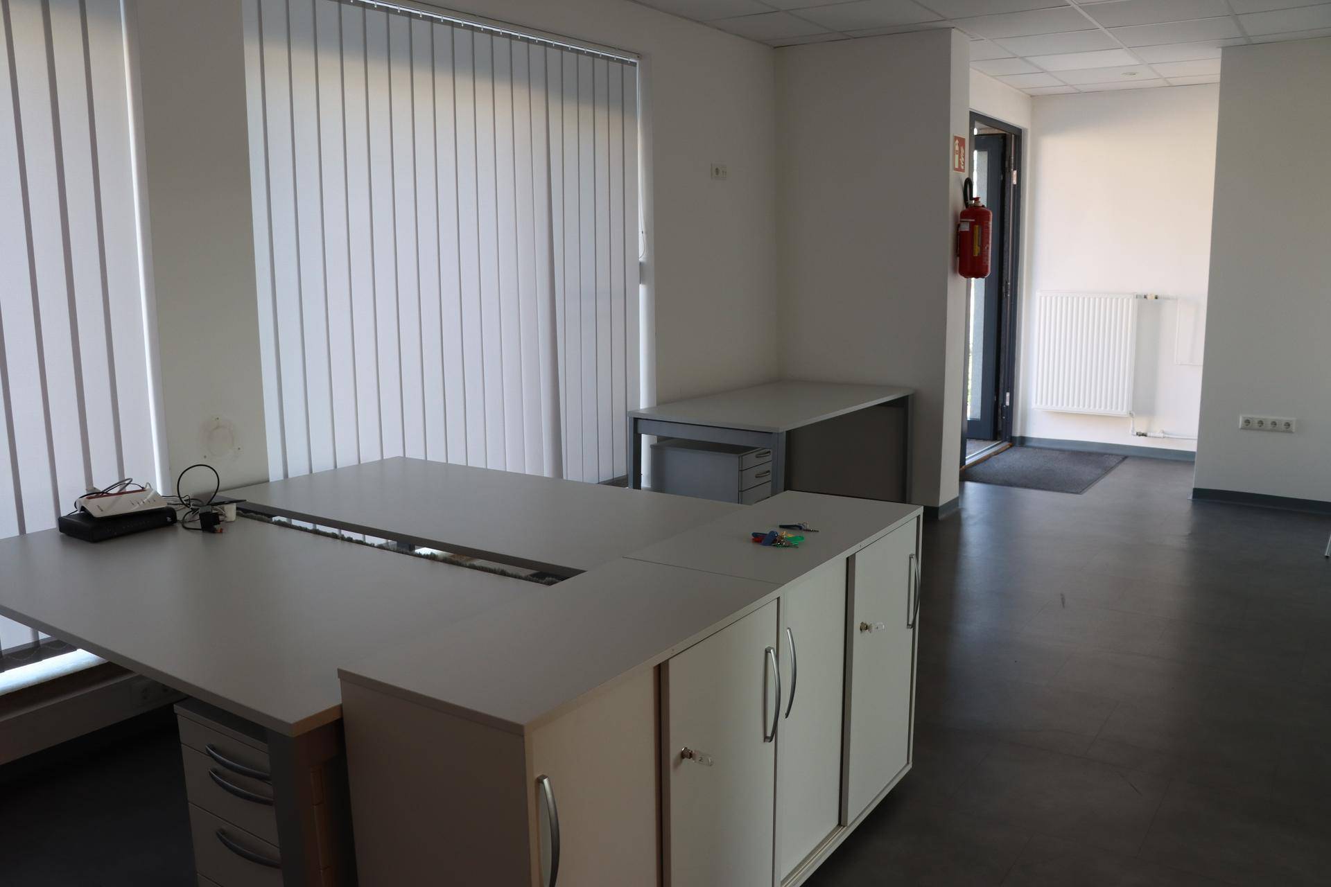 Büroarbeitsplatz mit Schreibtisch, Rollcontainer und vertikalen Jalousien an den Fenstern für Sichtschutz.