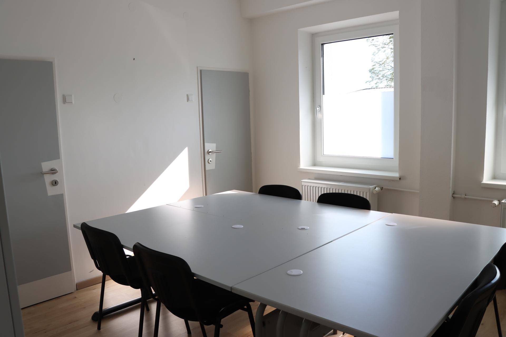 Konferenzraum mit großem modularem Tisch, Stühlen, hellen Wänden und Fenstern für Tageslicht.