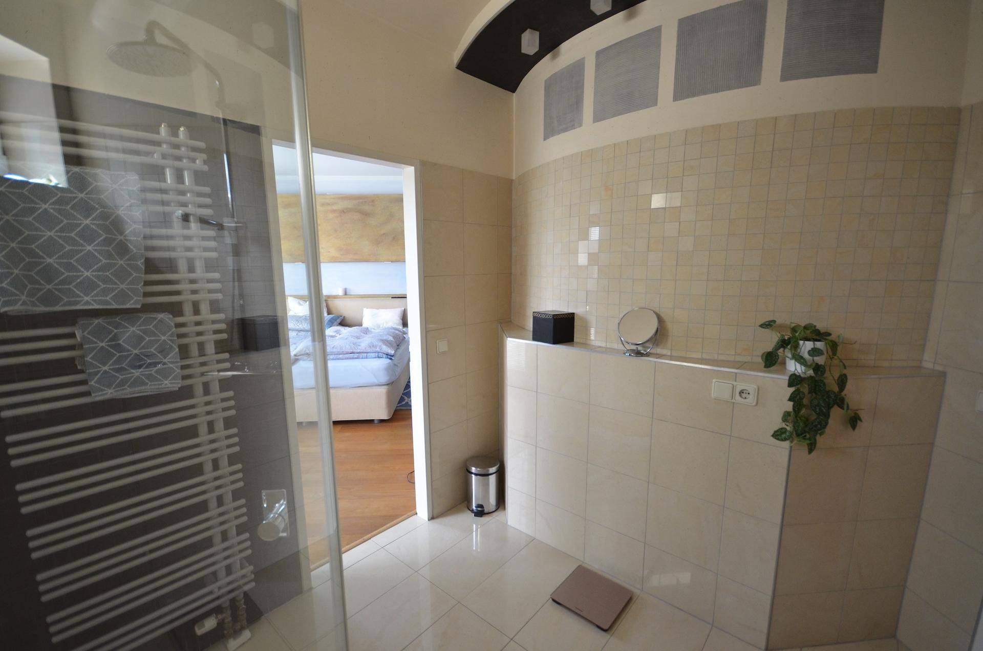 Gefliestes Badezimmer mit Spiegel und direktem Blick in das angrenzende Schlafzimmer.