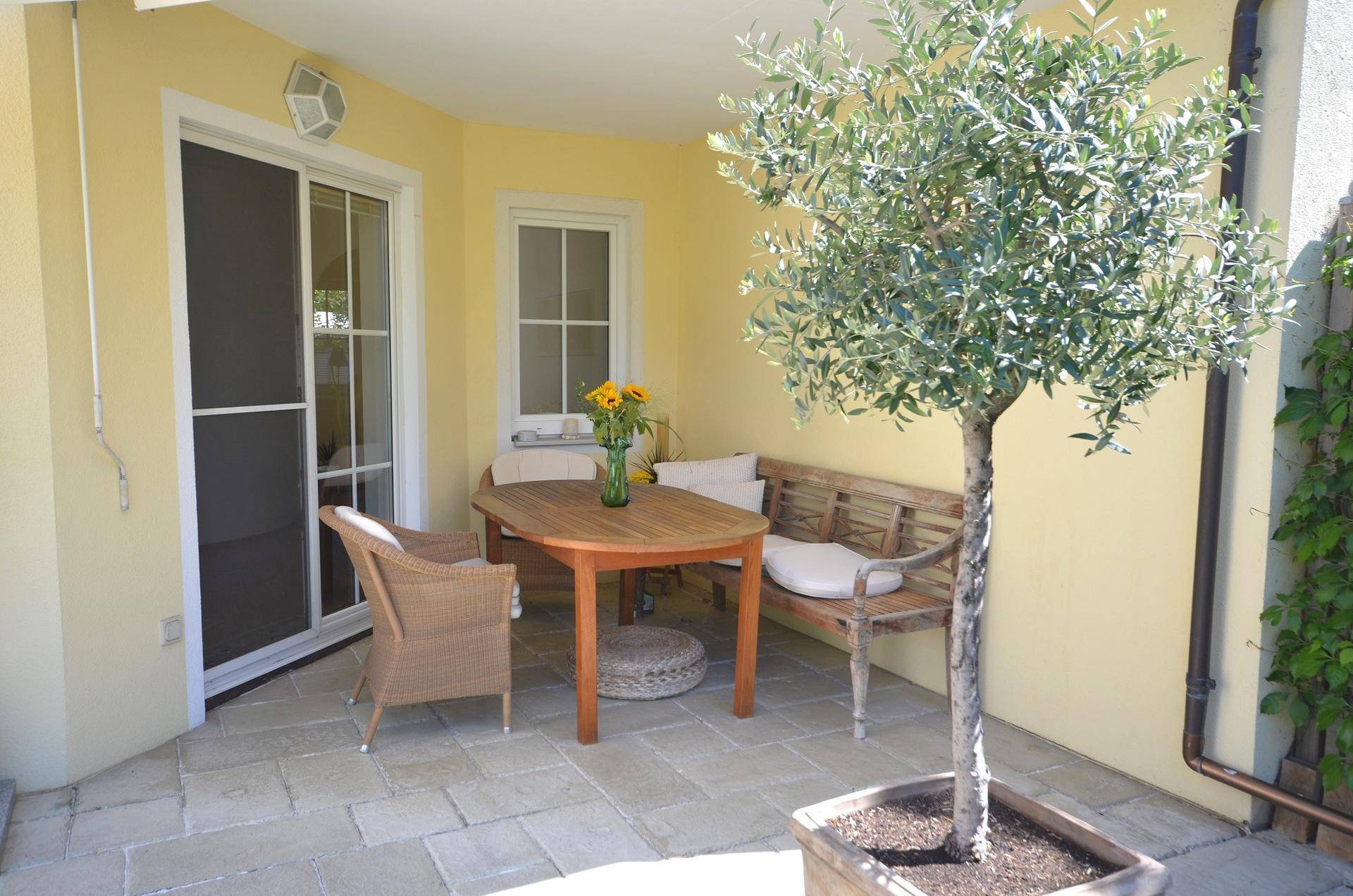 Gemütliche Terrasse mit Holztisch, Sitzgelegenheiten und einem Olivenbaum in einem Topf.