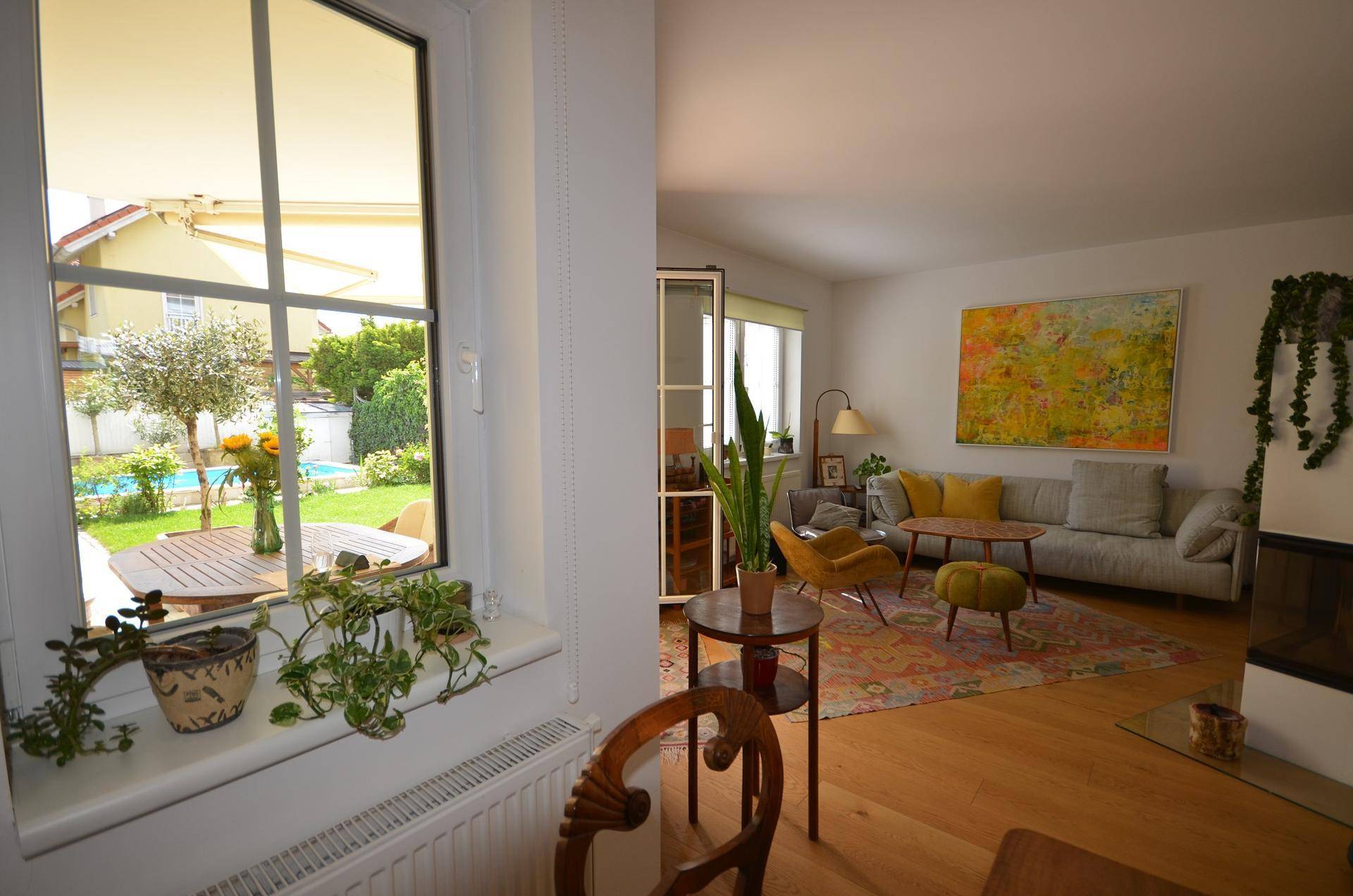 Blick aus dem Wohnzimmerfenster auf den grünen Garten mit Swimmingpool und Terrasse.
