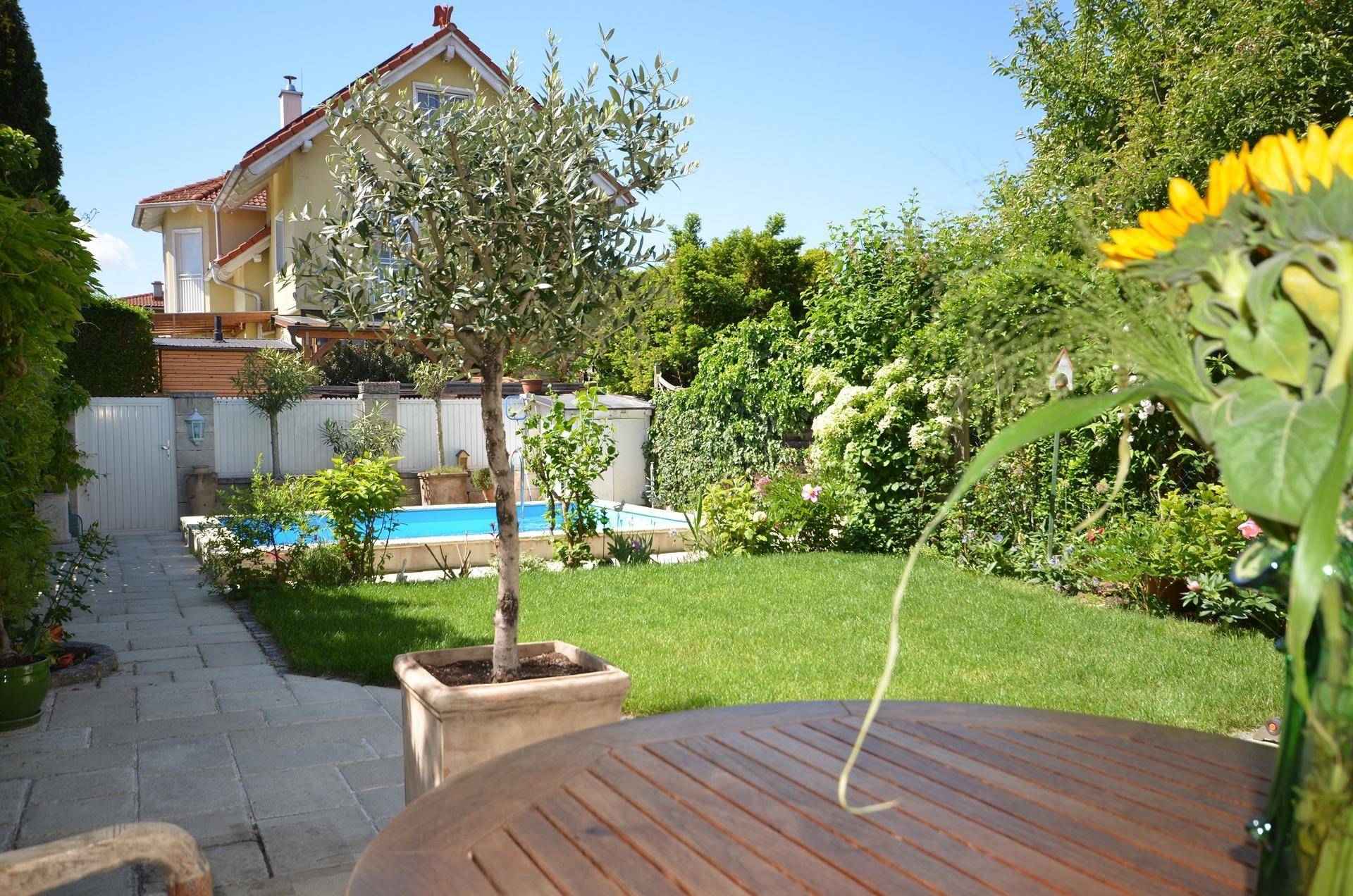 Blick vom Gartentisch auf den Swimmingpool, die Rasenfläche und das charmante Haus im Hintergrund.