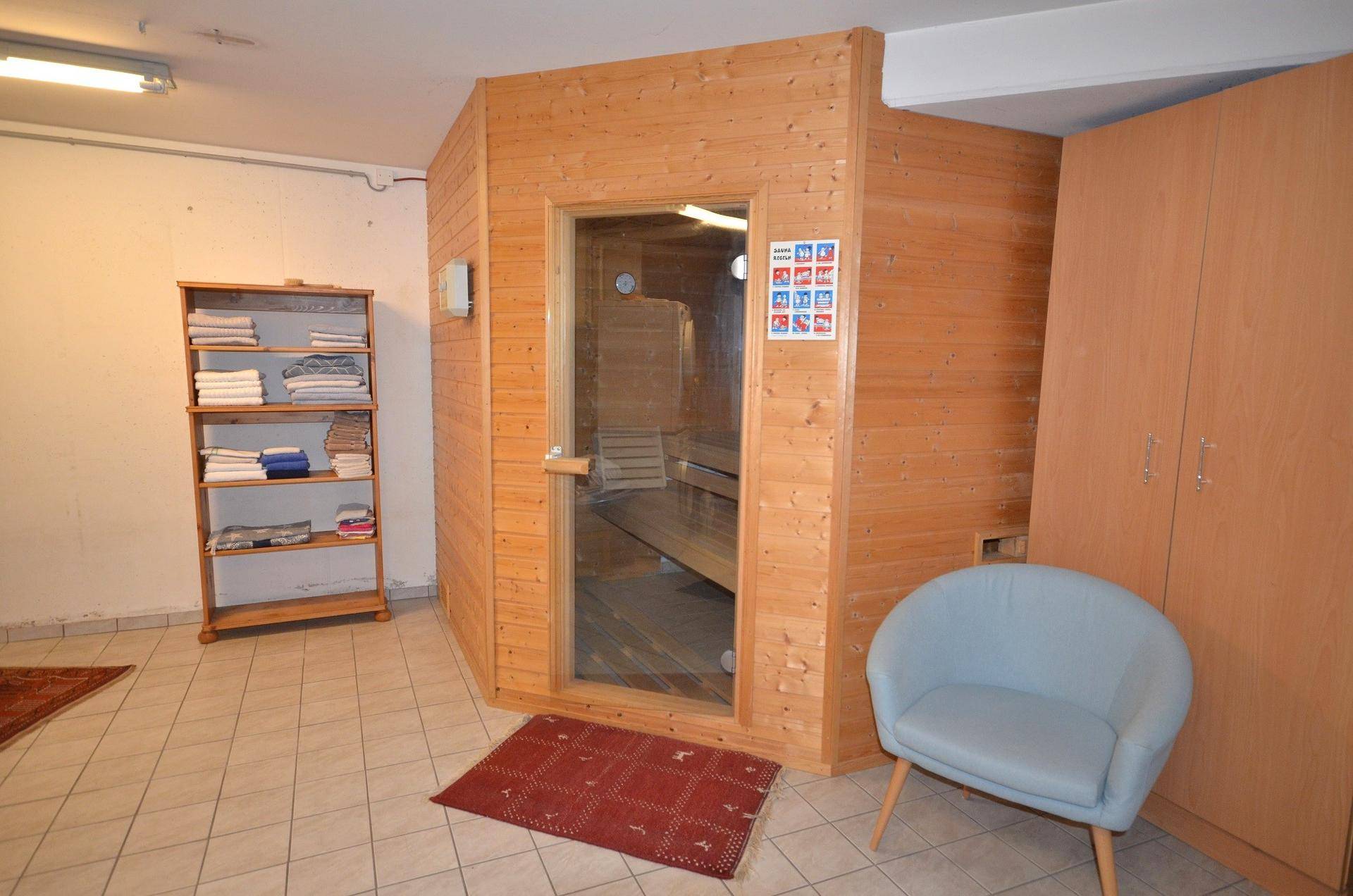Holzsauna mit Glastür in einem gefliesten Raum, ergänzt durch ein Regal und einen Sessel.