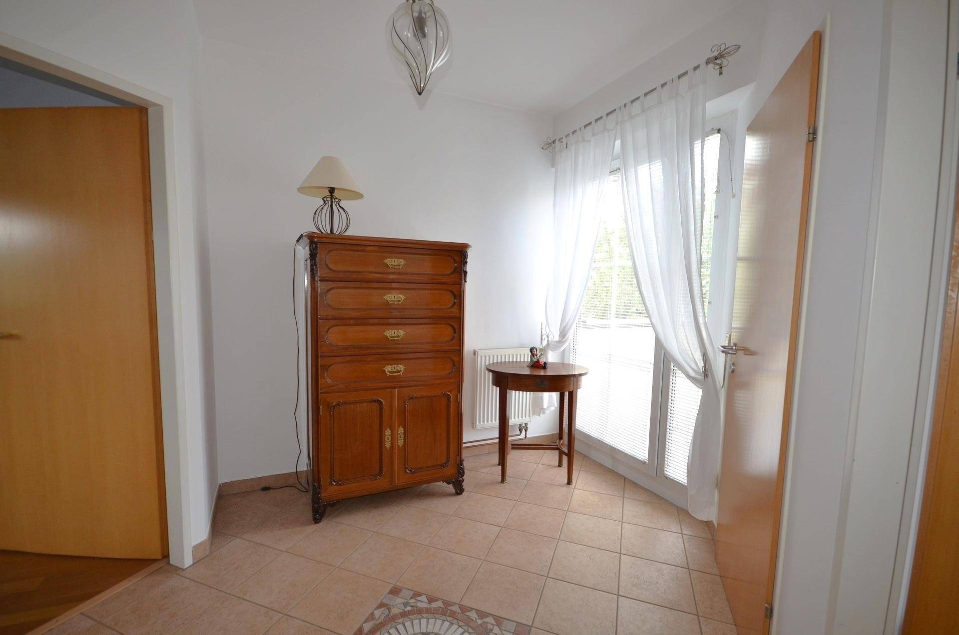 Heller Flur mit Fliesenboden, antiker Kommode und Fenster mit leichten Vorhängen.