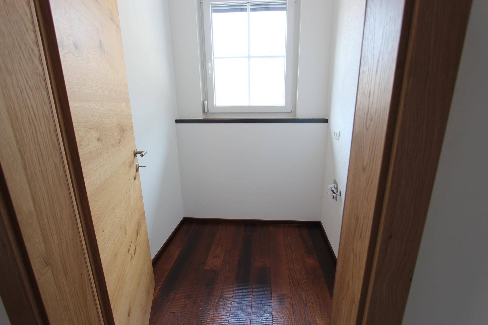 Kleiner, leerer Raum mit dunklem Holzboden und Fenster, ideal als Abstell- oder Hauswirtschaftsraum.