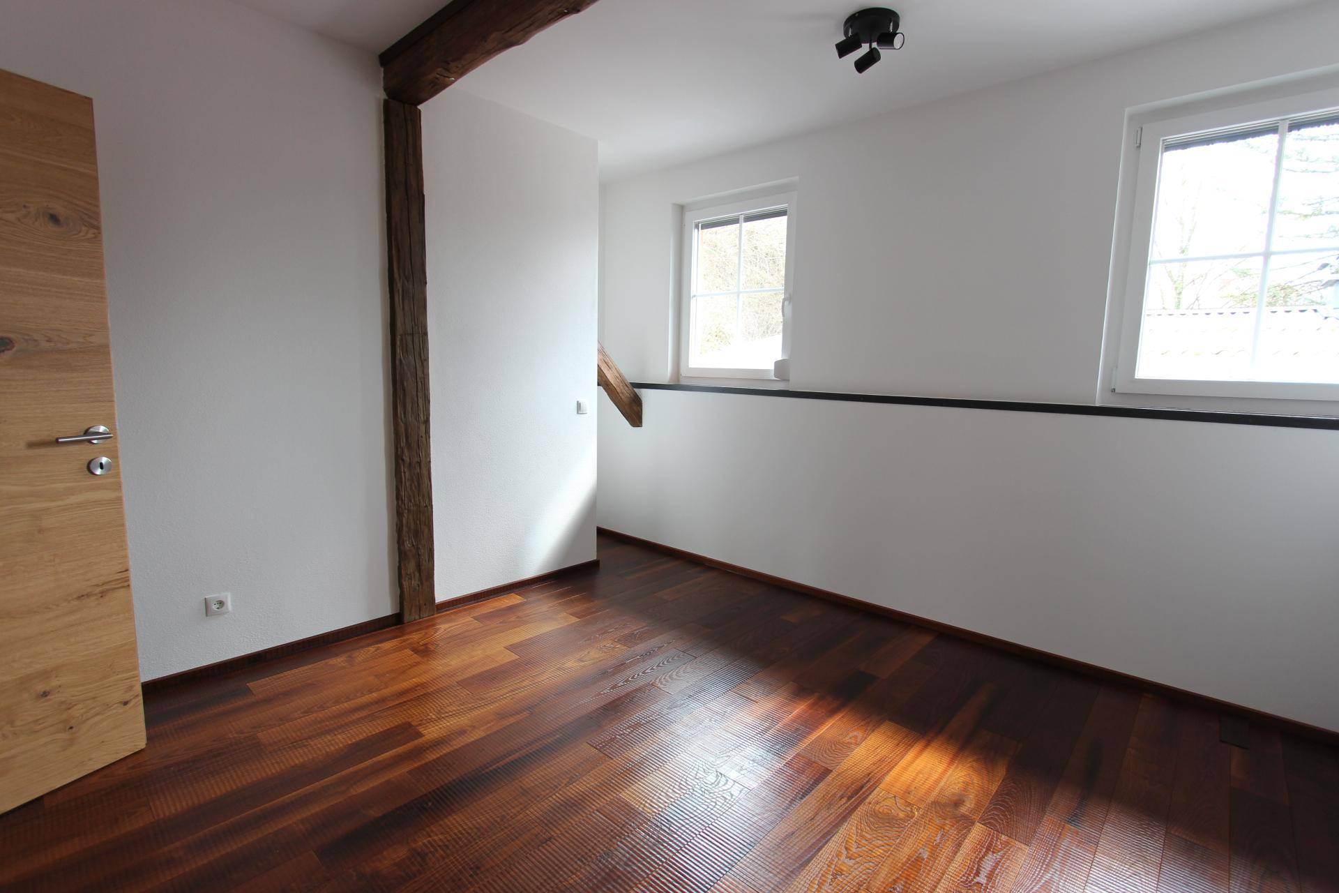 Leeres Schlafzimmer mit dunklem Holzboden, weißen Wänden und zwei Fenstern, durch die Licht einfällt.