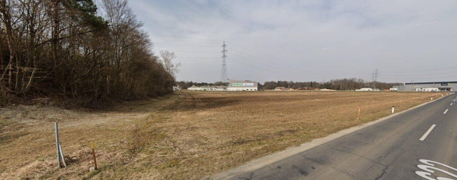 Weitläufiges Feld neben einer Straße mit Waldrand und Industriegebäuden im Hintergrund.