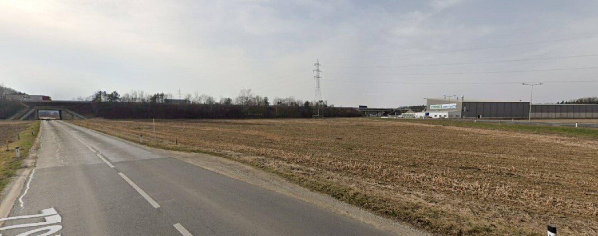 Blick über ein weites Feld entlang einer Straße mit einer Autobahnbrücke im Hintergrund.