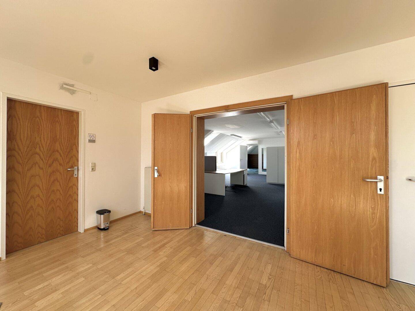 Heller Büroraum mit Holzboden und zwei Holztüren, die zu weiteren Bereichen führen.