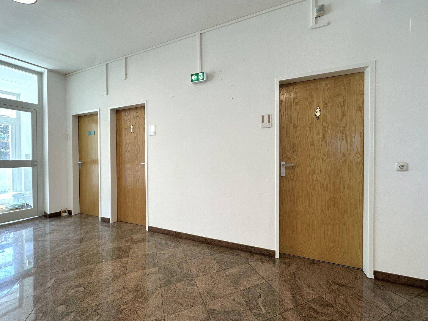 Flurbereich mit Marmorboden und drei Holztüren, darunter zwei Toiletten mit Piktogrammen.