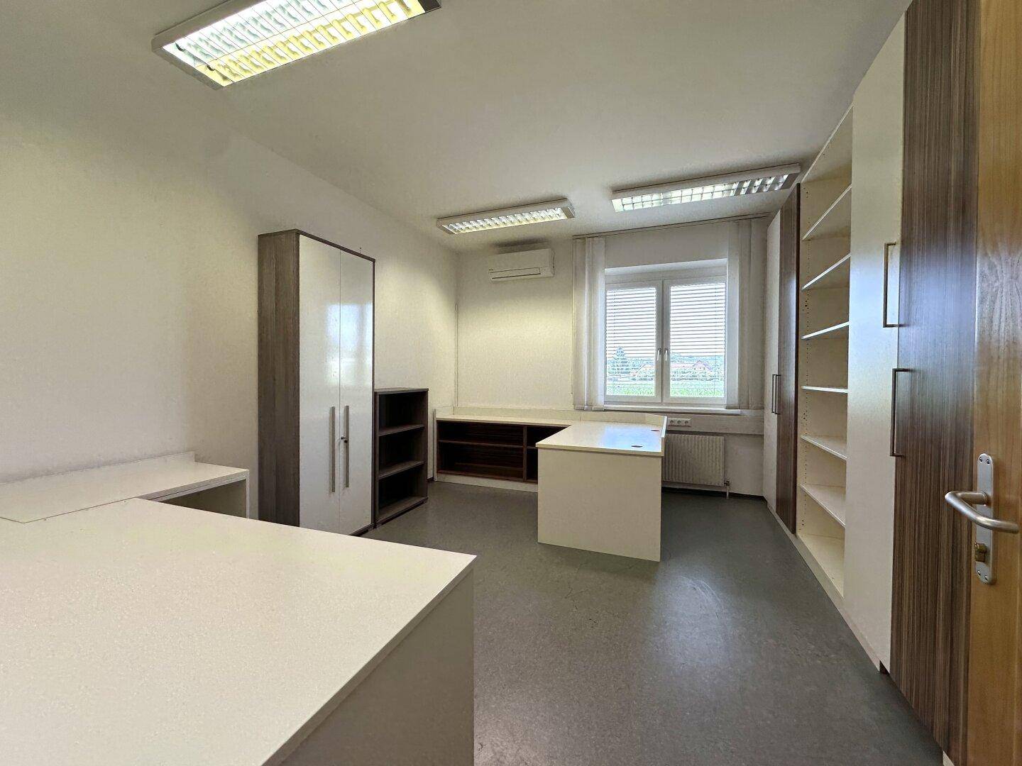 Helles Büro mit Schränken, einem Schreibtisch und einem Fenster mit Blick ins Grüne.