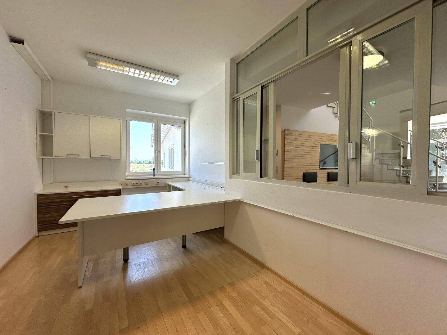 Büroraum mit Holzboden und einem großen Fenster, das einen Blick in den Hauptflur ermöglicht.