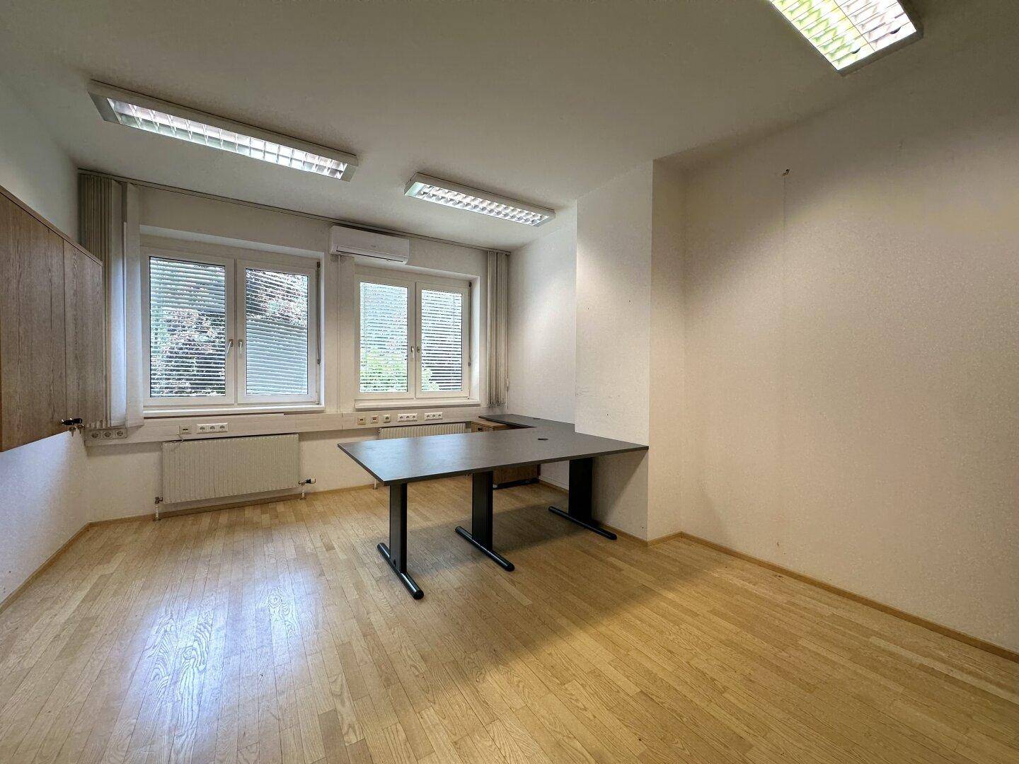 Büro mit Holzfußboden, dunklem Schreibtisch und Fenstern mit Jalousien für angenehmes Licht.
