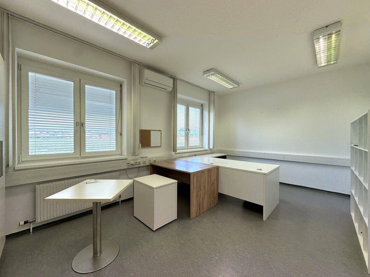 Helles Büro mit grauem Vinylboden, mehreren Schreibtischen und Fenstern mit Jalousien.