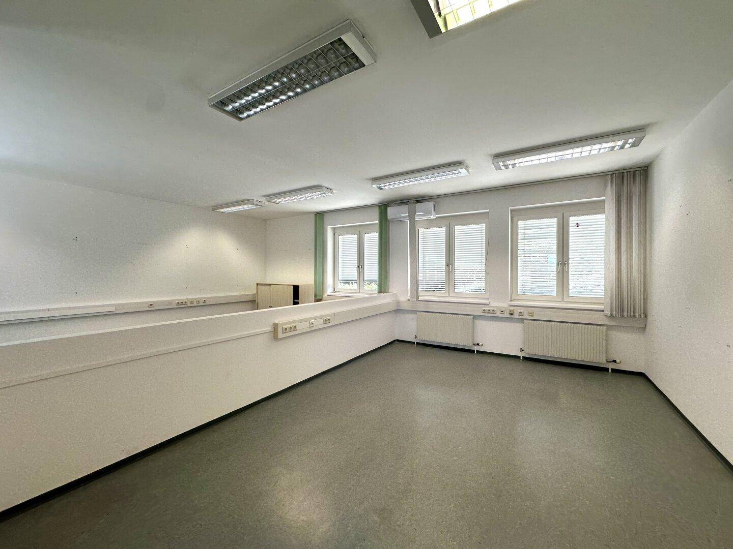 Geräumiges Büro mit grauem Vinylboden, Fenstern und einer niedrigen Trennwand für offene Arbeitsbereiche.
