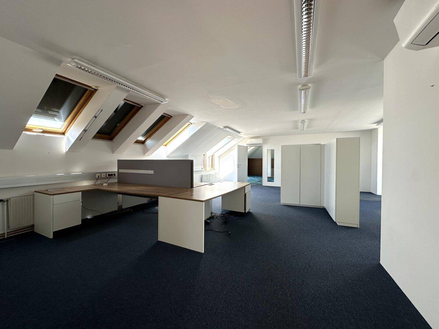 Geräumiges Dachgeschossbüro mit dunklem Teppichboden, Schreibtischen und mehreren Dachfenstern.
