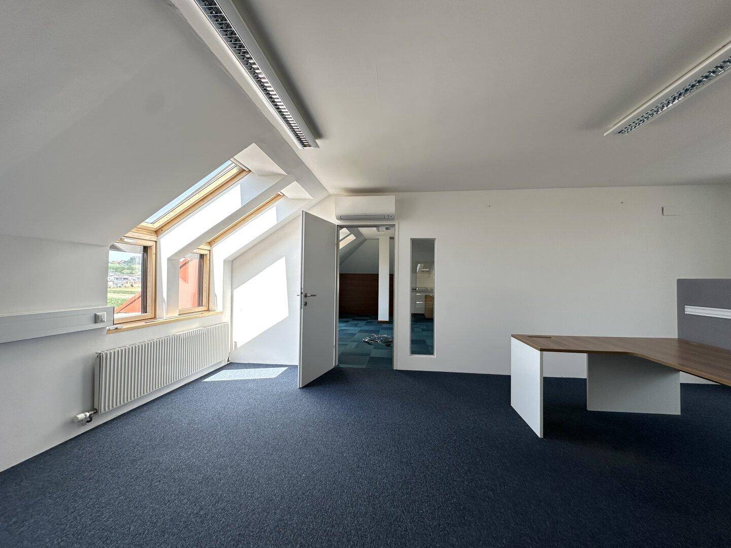 Helles Dachgeschossbüro mit Teppichboden, Heizkörper und offener Tür zu einem angrenzenden Bereich.