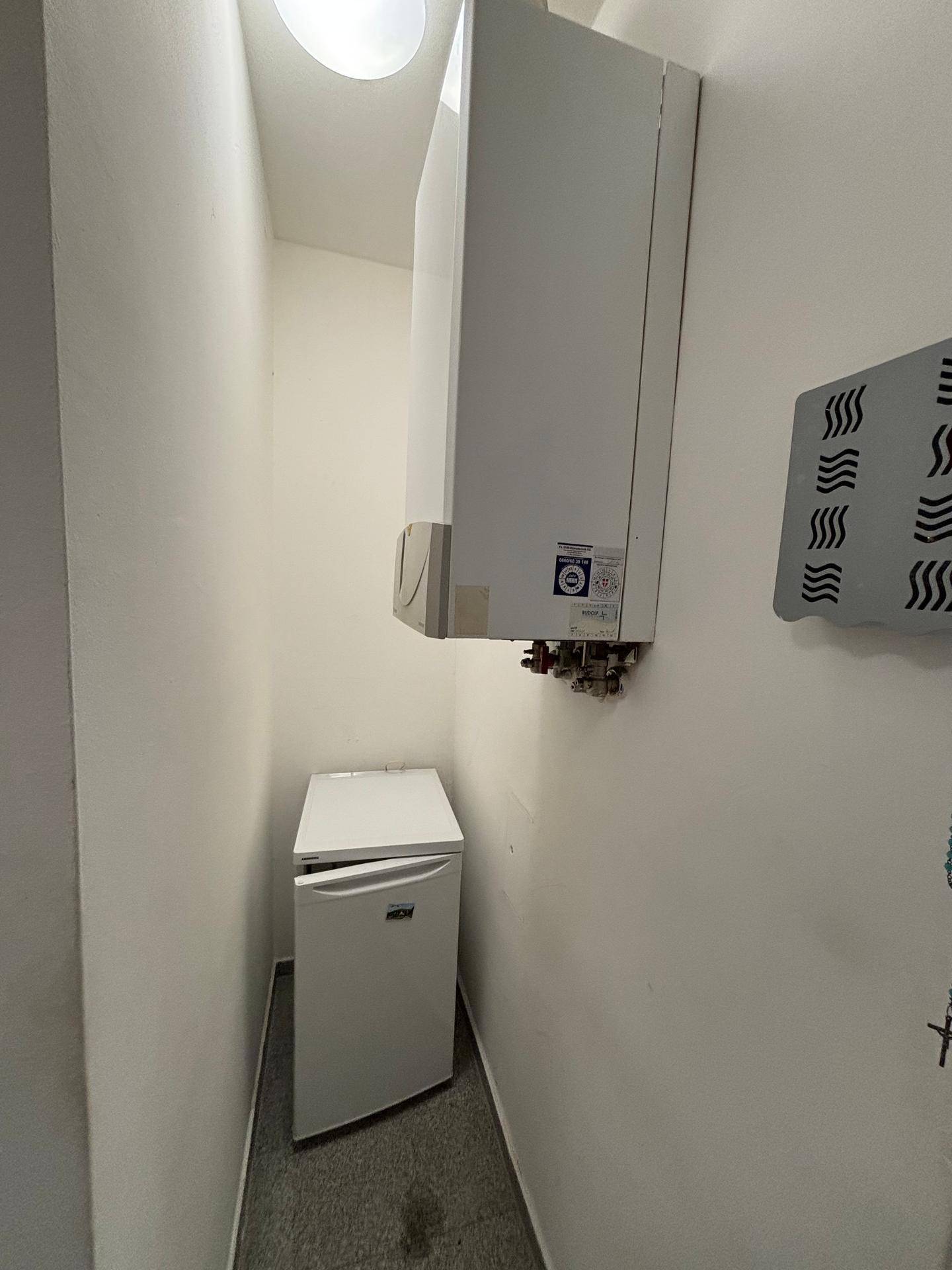Hauswirtschaftsraum mit wandmontiertem Boiler, kleinem Kühlschrank und grauem Fliesenboden.