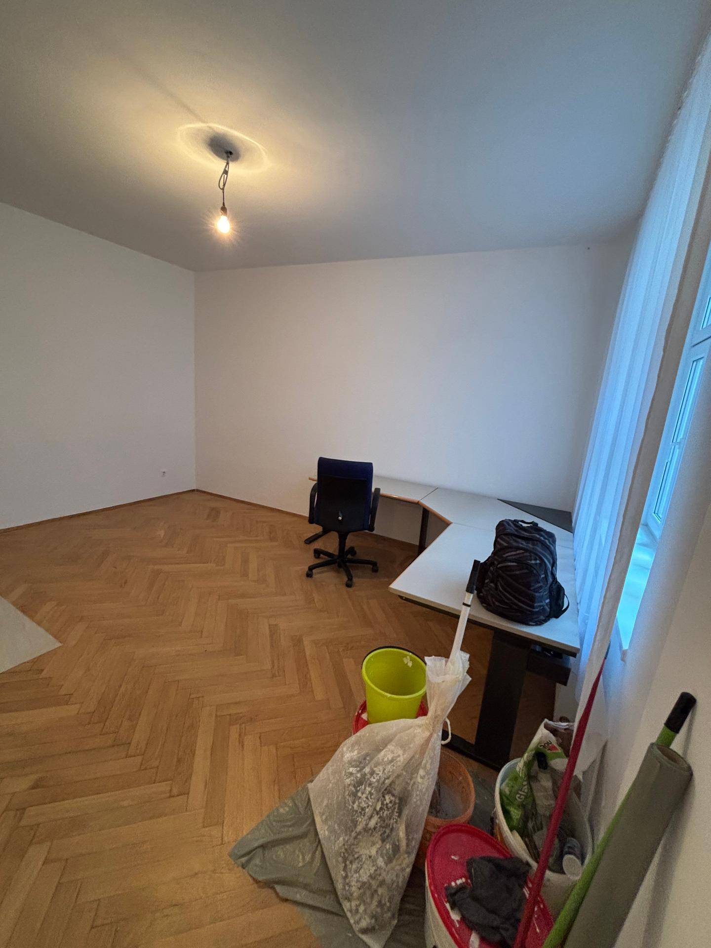 Heller Raum mit Fischgrätparkett und großem Fenster, derzeit als Arbeitsbereich genutzt mit Reinigungsutensilien.