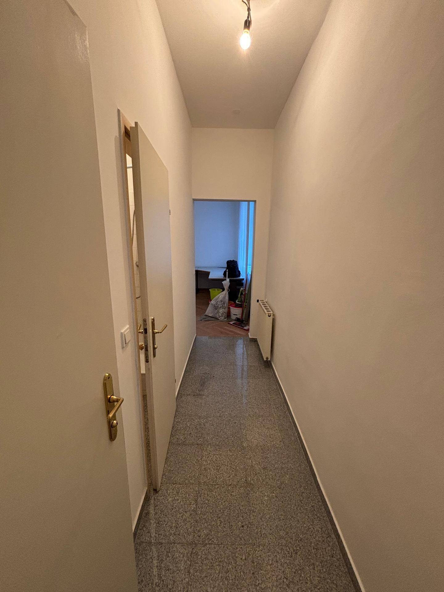 Schmaler Flur mit grauem Fliesenboden und Heizkörper, der den Blick in einen angrenzenden Raum freigibt.