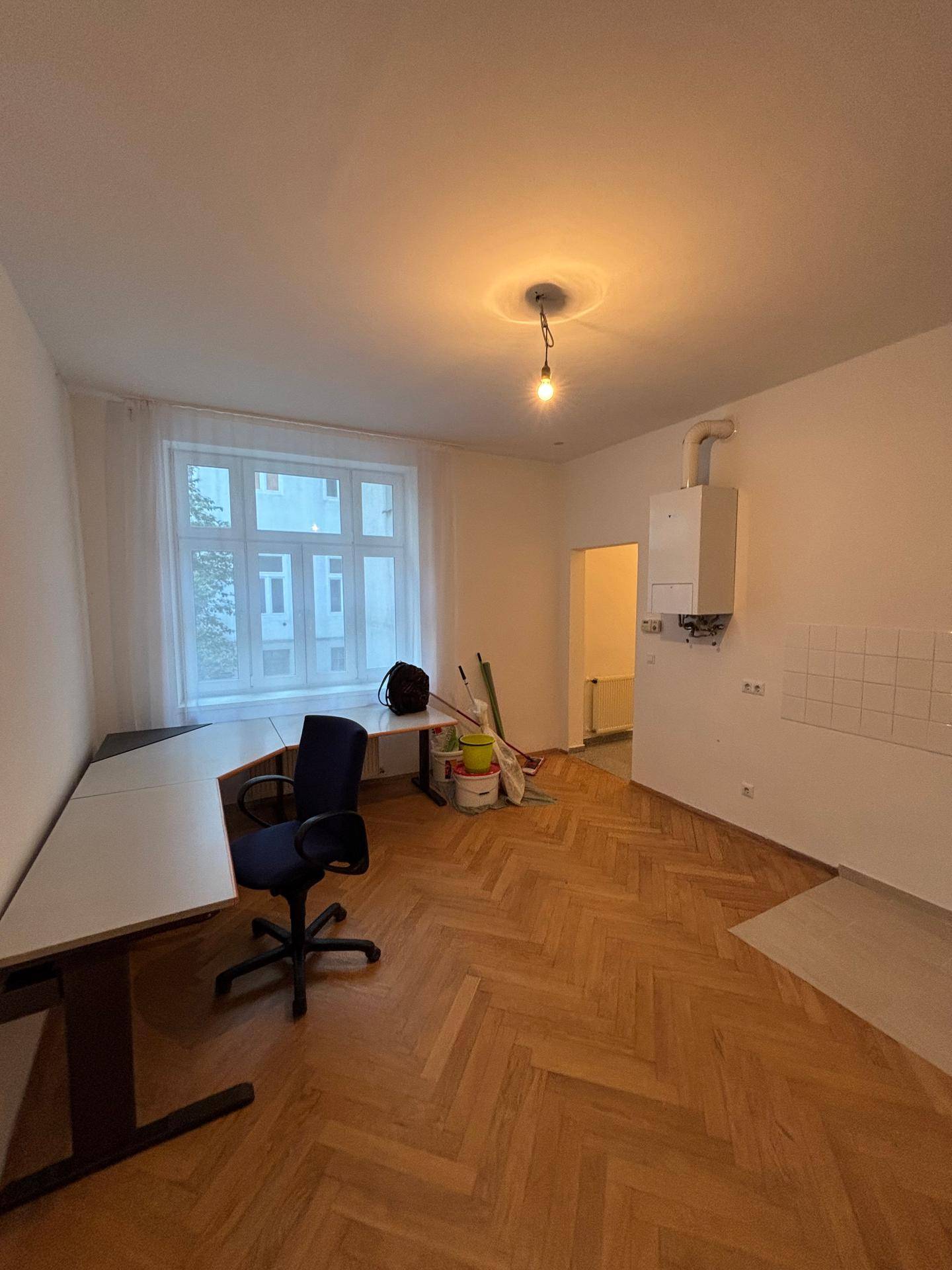 Geräumiger Wohnbereich mit Fischgrätparkett, großem Fenster und integrierter Küchenzeile.