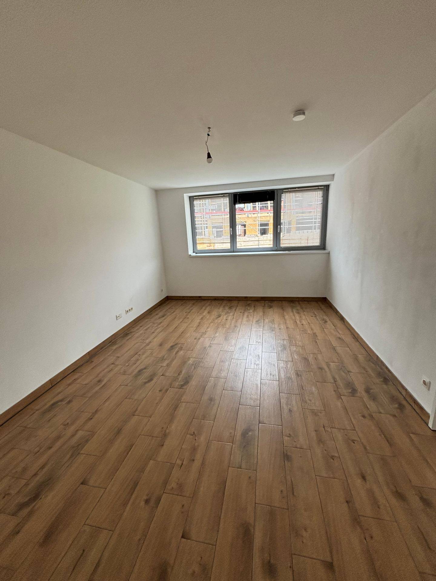 Geräumiger, leerer Raum mit Holzboden und einem großen Fenster, das viel Tageslicht hereinlässt.