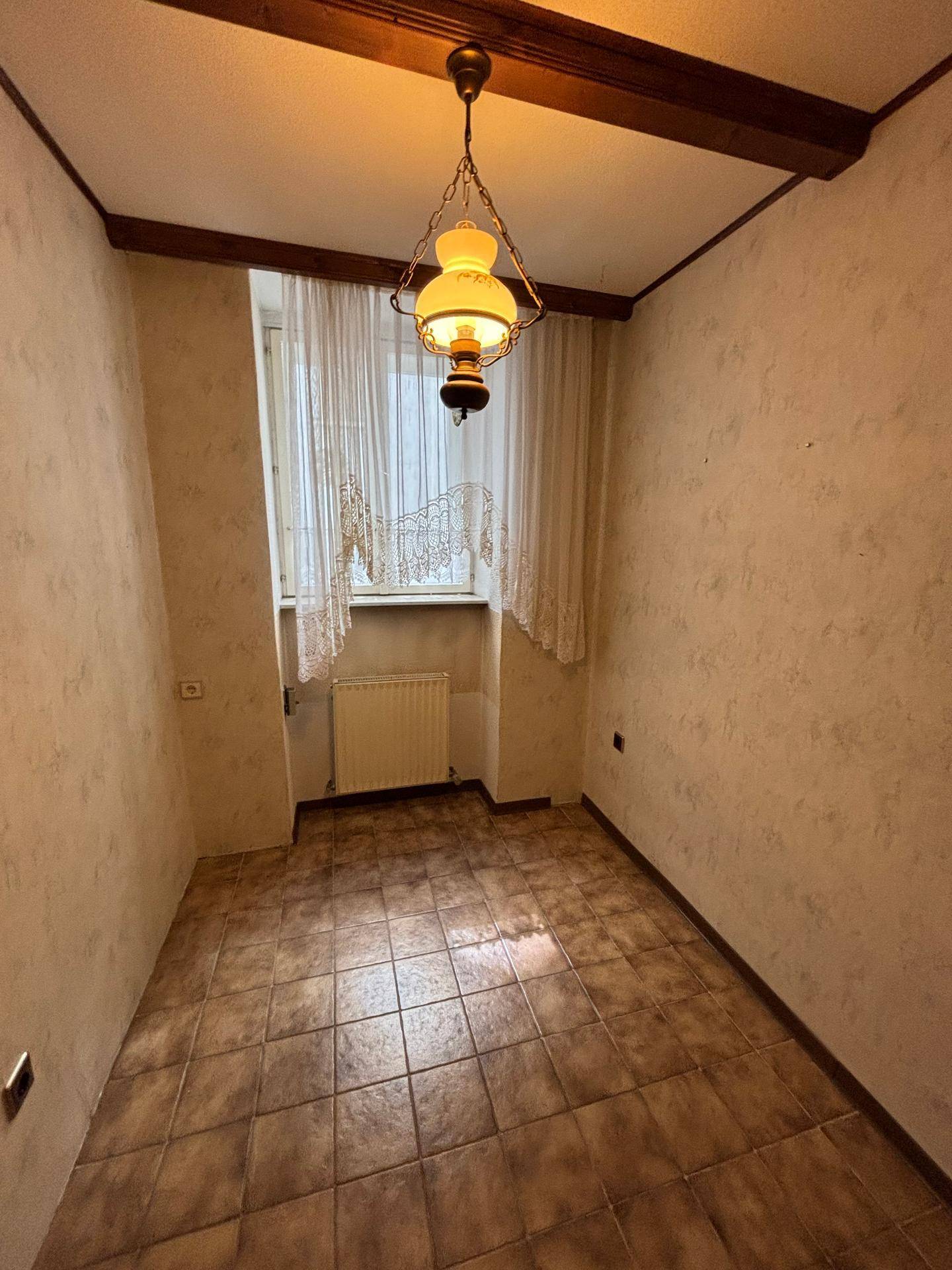 Helles Zimmer mit Fliesenboden, Holzbalken und einem Fenster mit traditionellen Vorhängen.