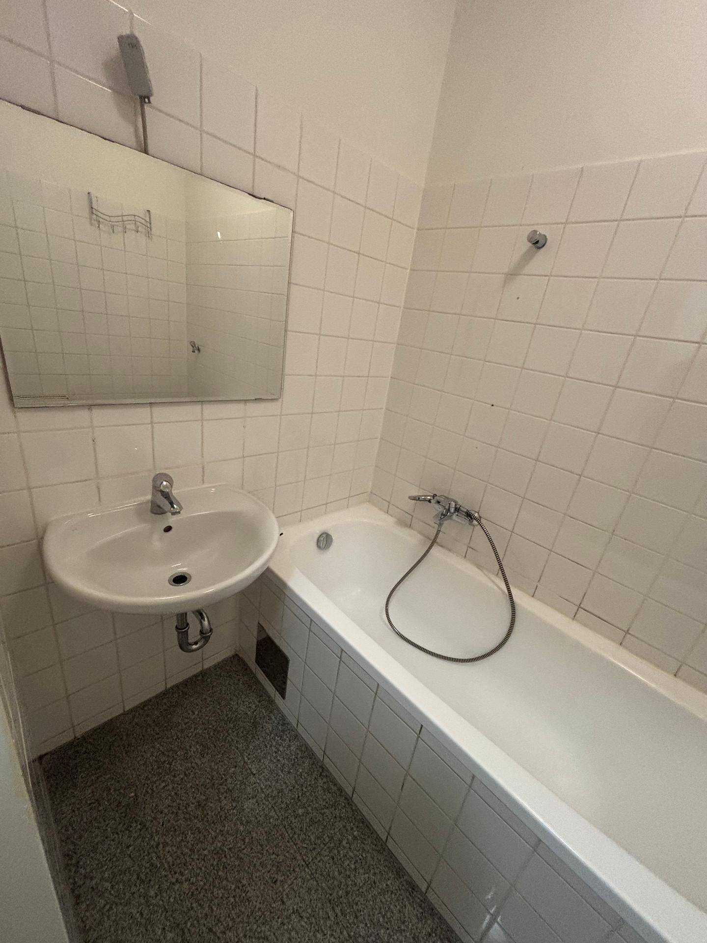 Badezimmer mit Badewanne, Waschbecken, Spiegel und weißen Wandfliesen auf grauem Fliesenboden.