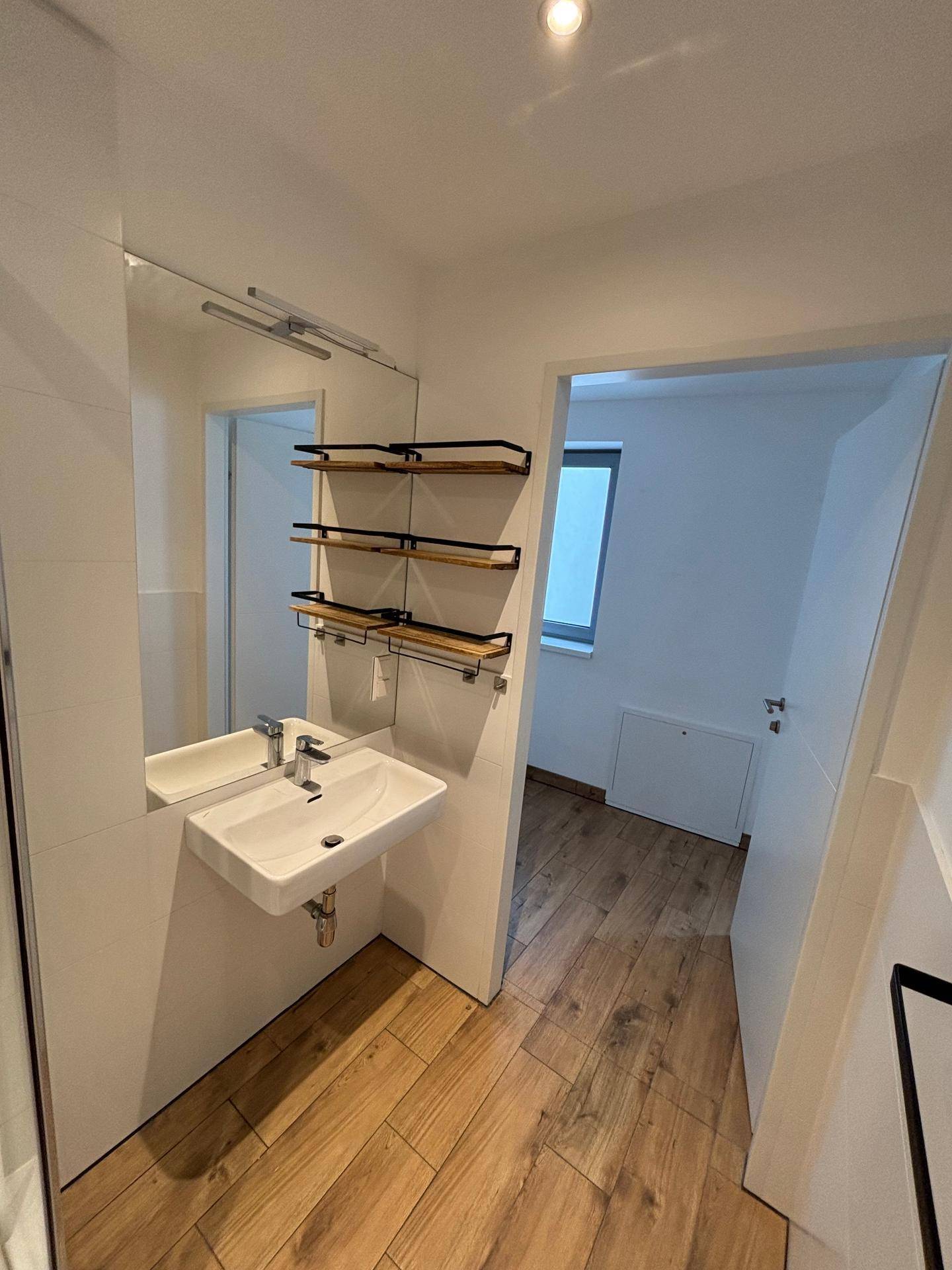 Badezimmer mit Waschbecken, großem Spiegel und praktischen Wandregalen, angrenzend an einen weiteren Raum.