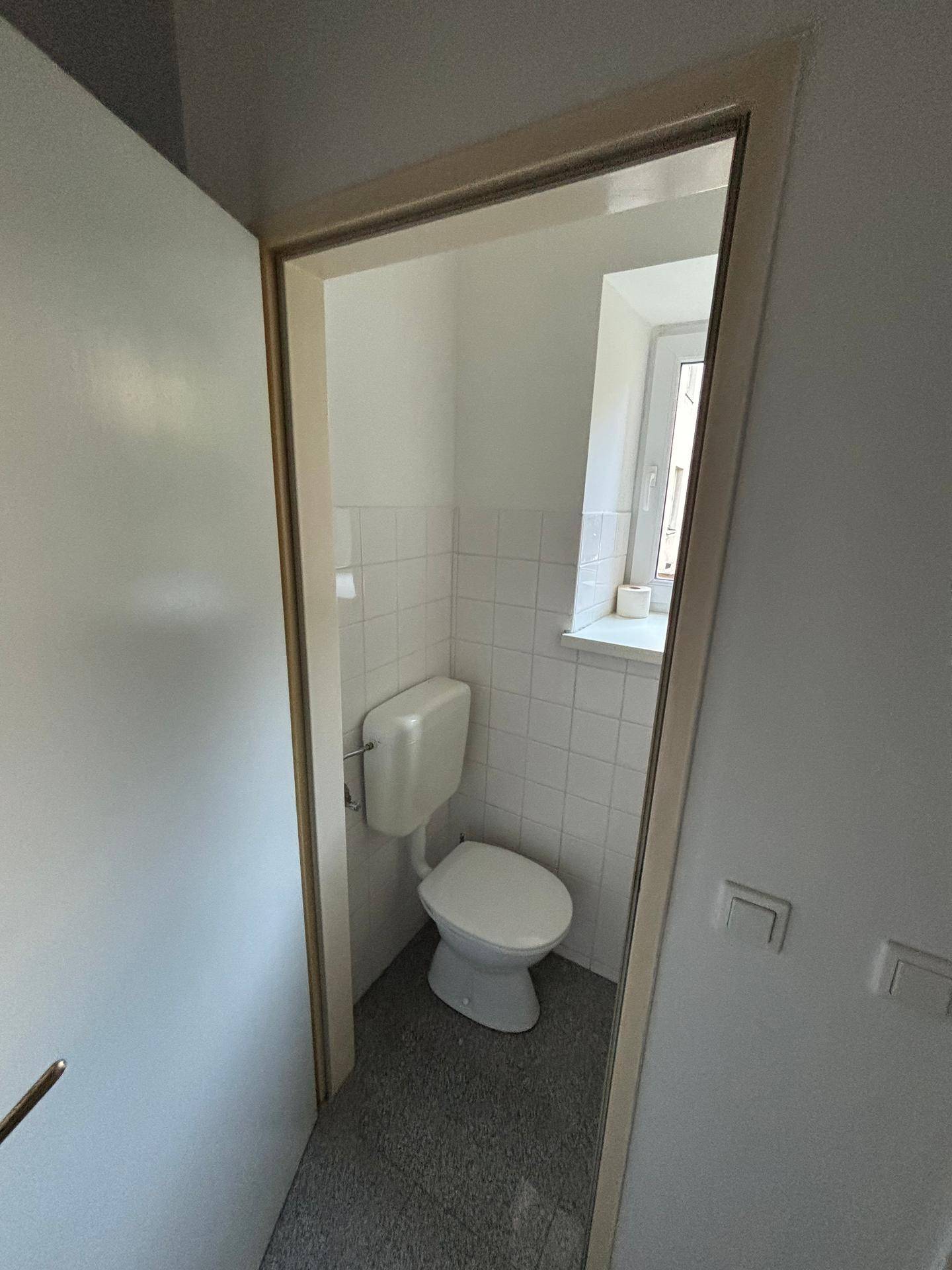 Kleiner Toilettenraum mit Fenster, weißer Keramik und weißen Wandfliesen über grauem Boden.