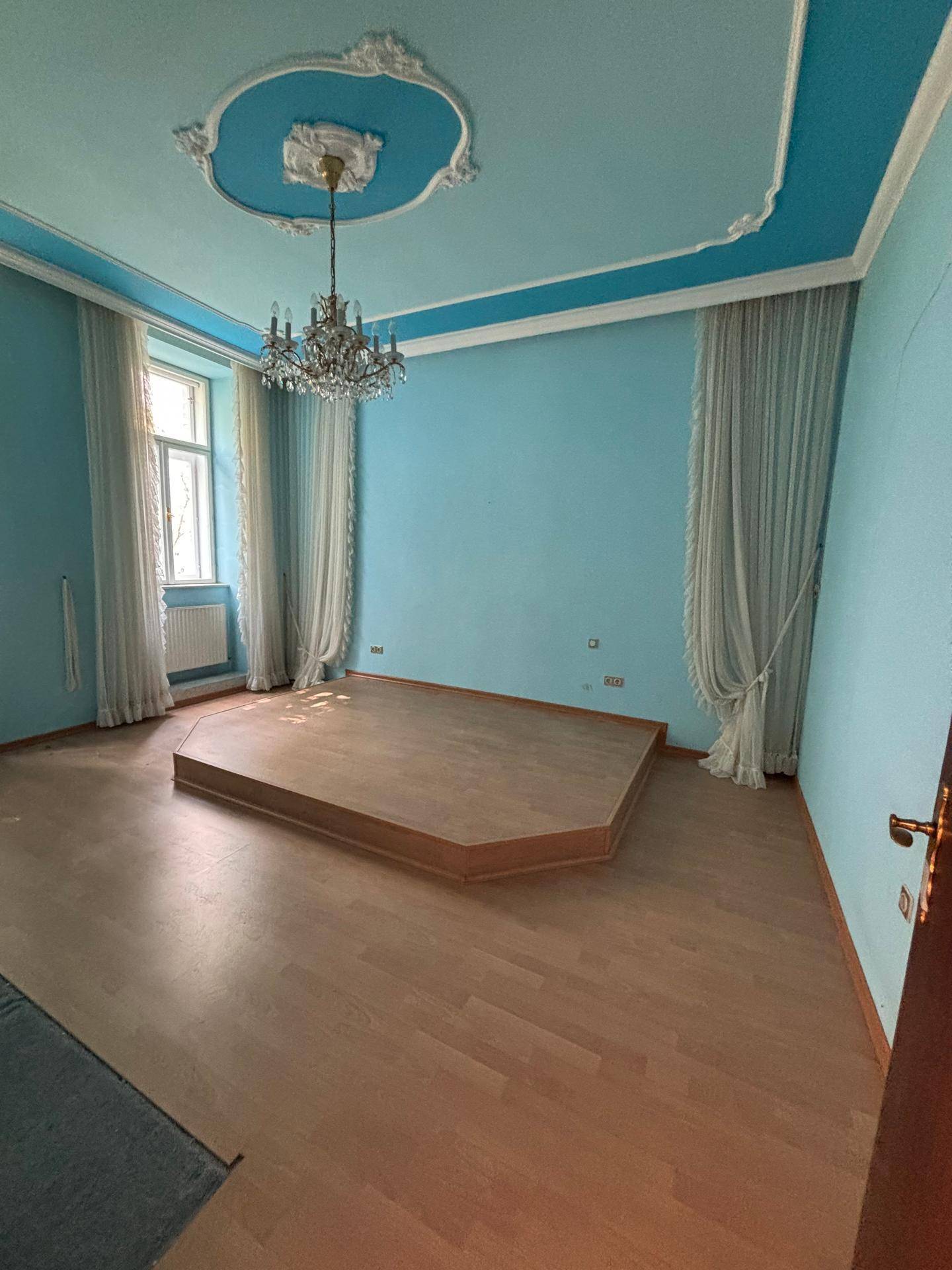 Helles Zimmer mit blauen Wänden, Parkettboden, einer erhöhten Plattform und Kronleuchter.