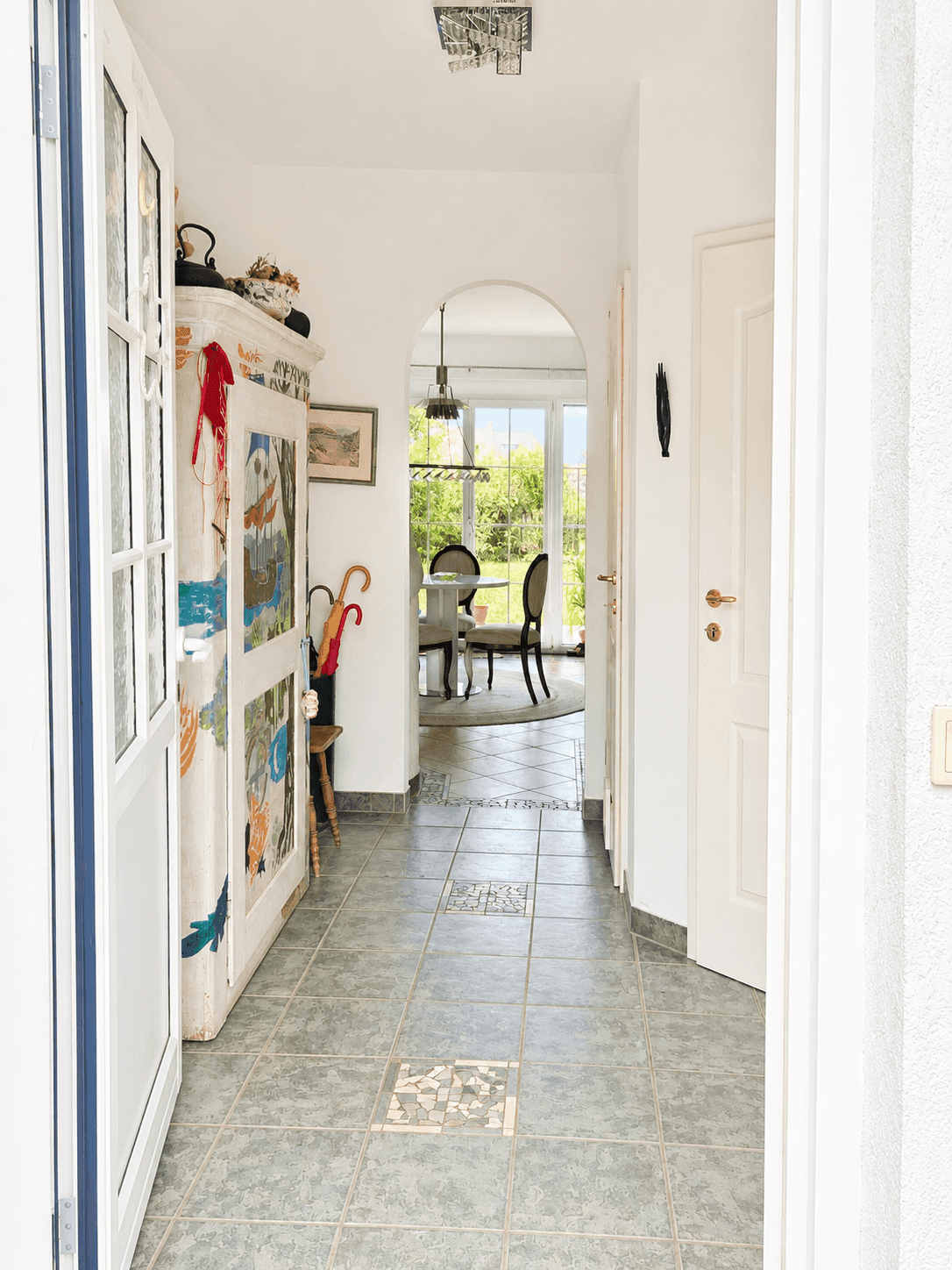 Heller Eingangsbereich mit Fliesenboden, farbenfrohem Schrank und Blick durch einen Bogen in den Essbereich mit Garten.