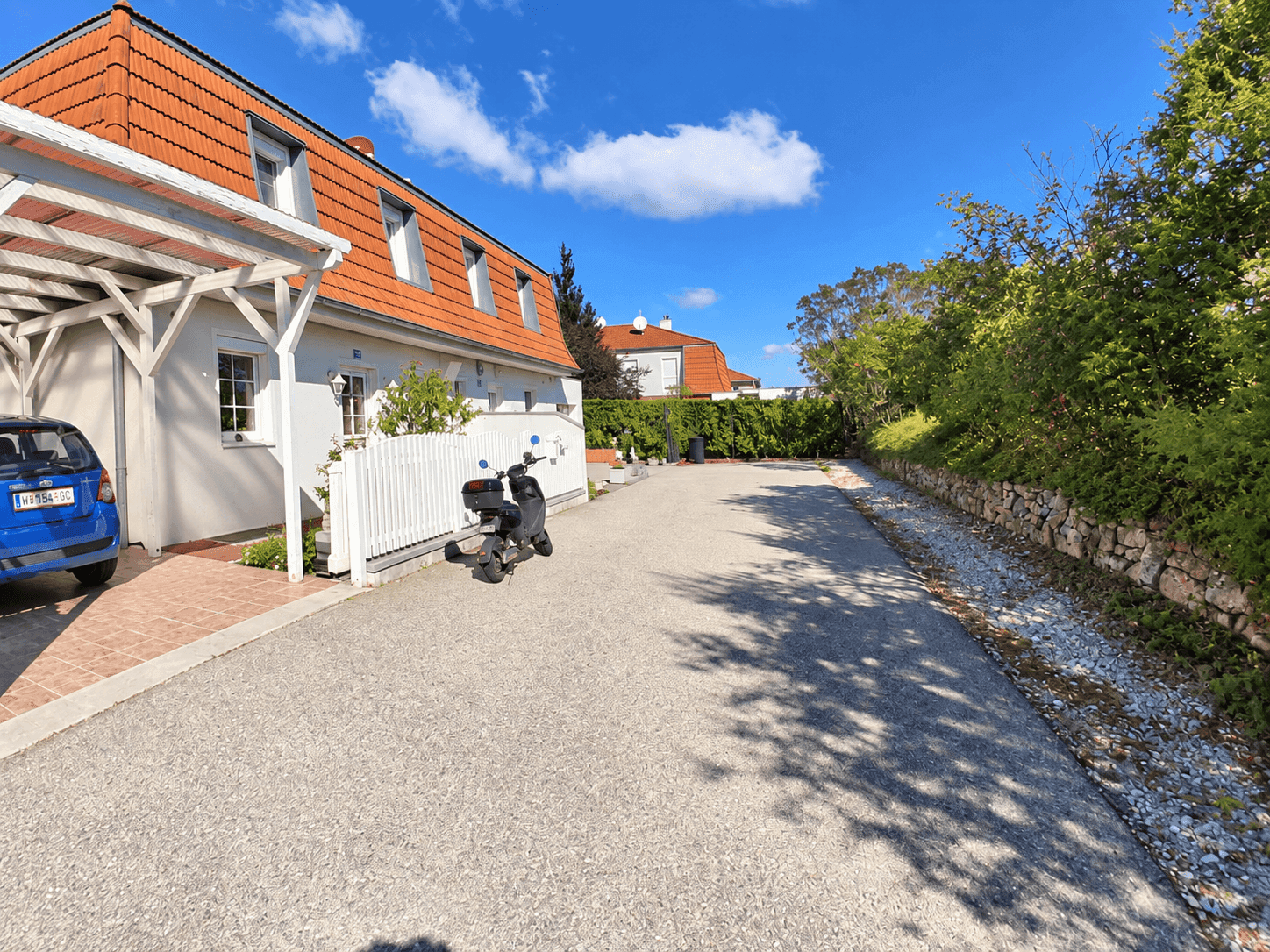 Seitenansicht eines Hauses mit rotem Ziegeldach, Schotterauffahrt und einem Carport für ein Fahrzeug.