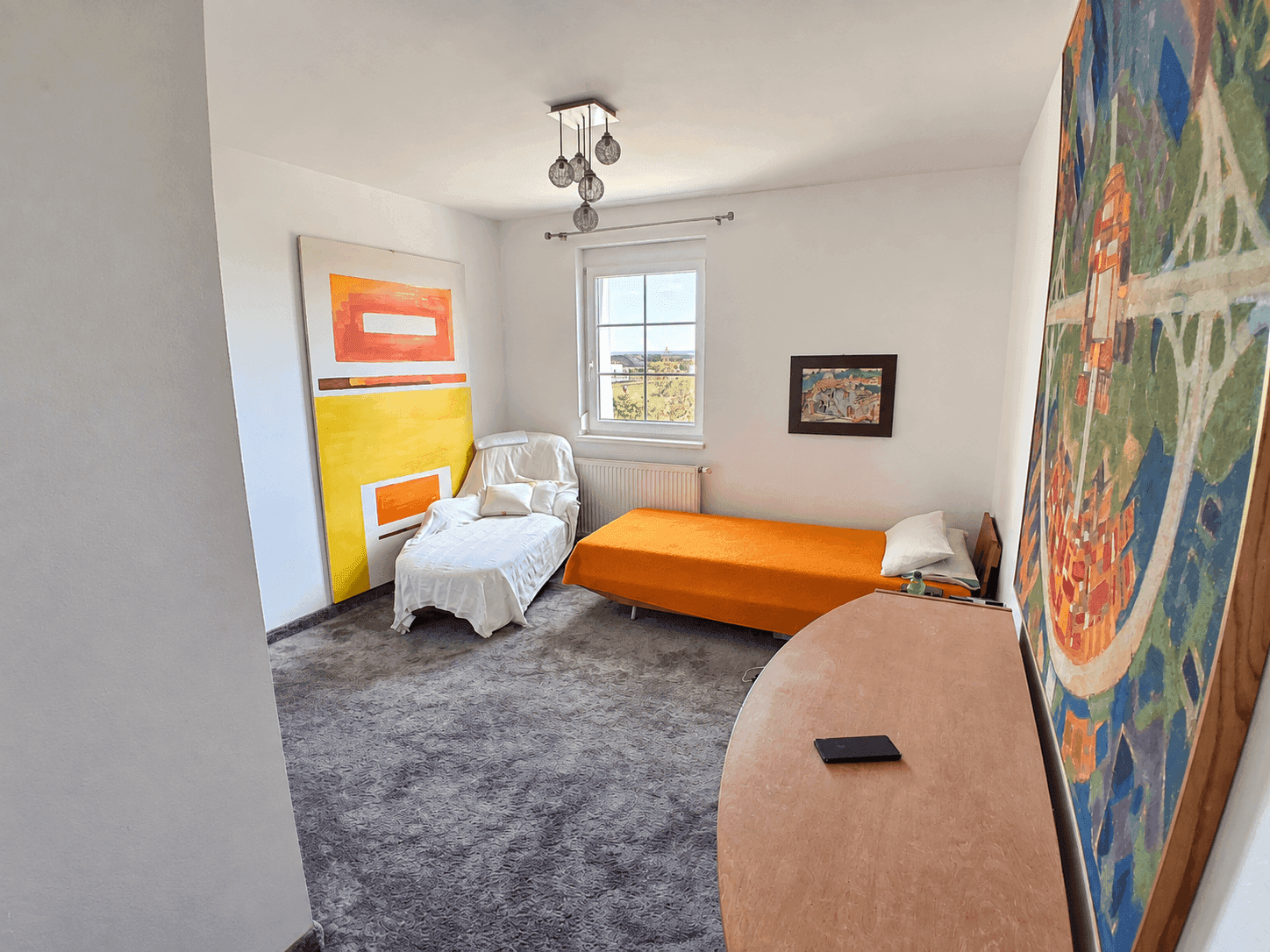 Helles Zimmer mit Teppichboden, Einzelbett und farbenfrohen Kunstwerken.