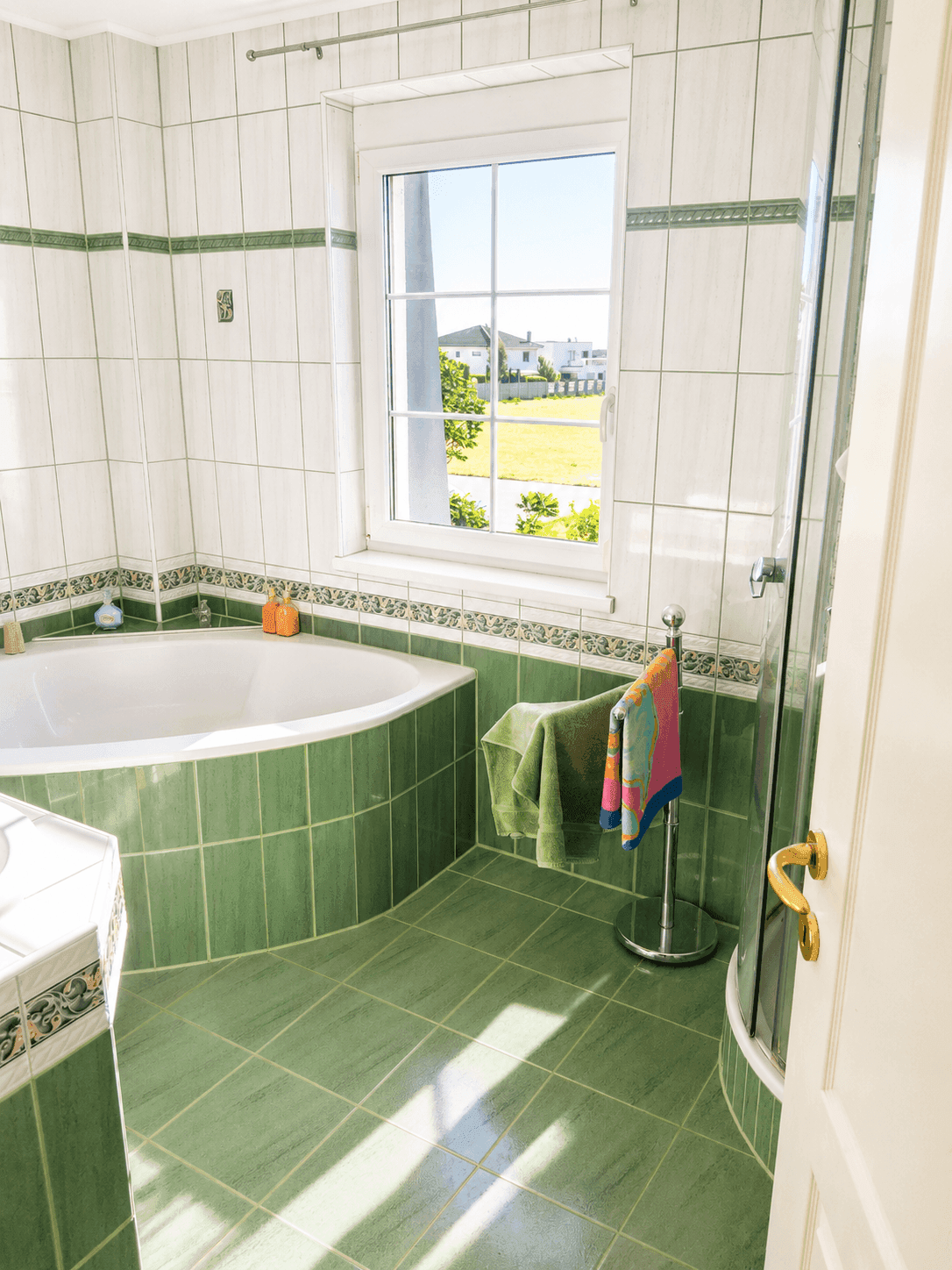Badezimmer mit grünen und weißen Fliesen, Eckbadewanne, Dusche und Fenster.